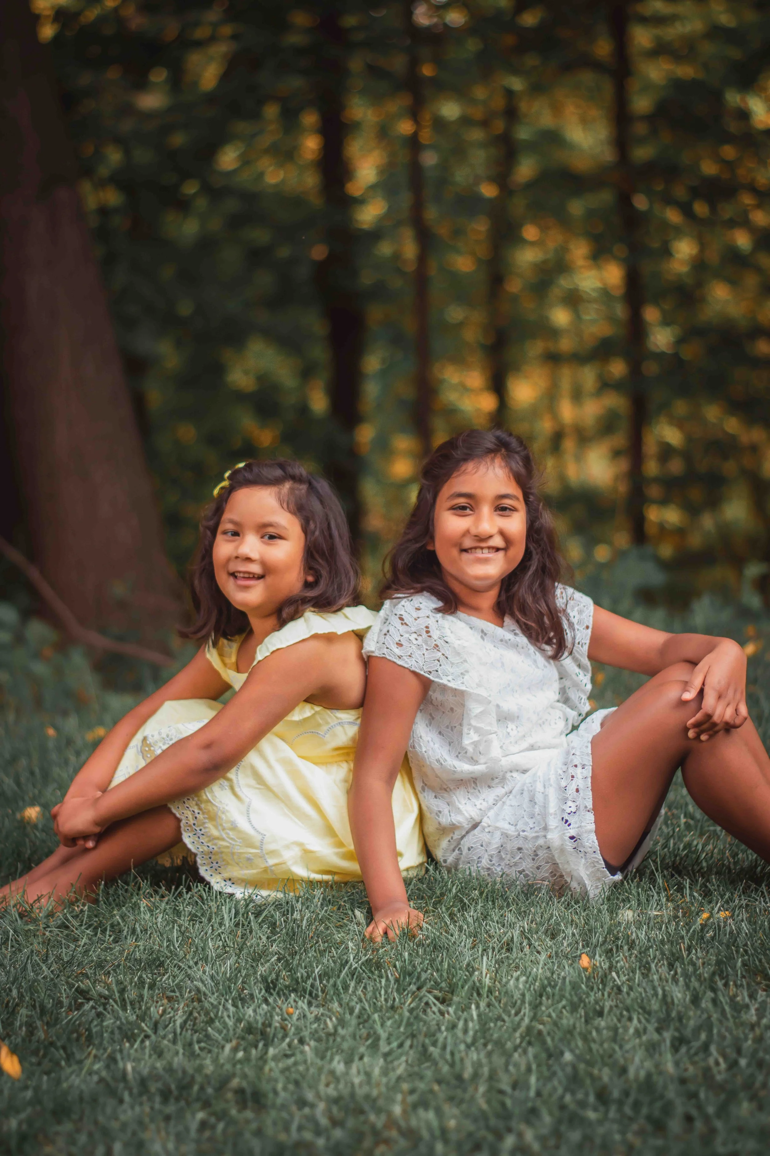 Two young girls sitting back-to-back on the grass in a wooded area, smiling at the camera.
