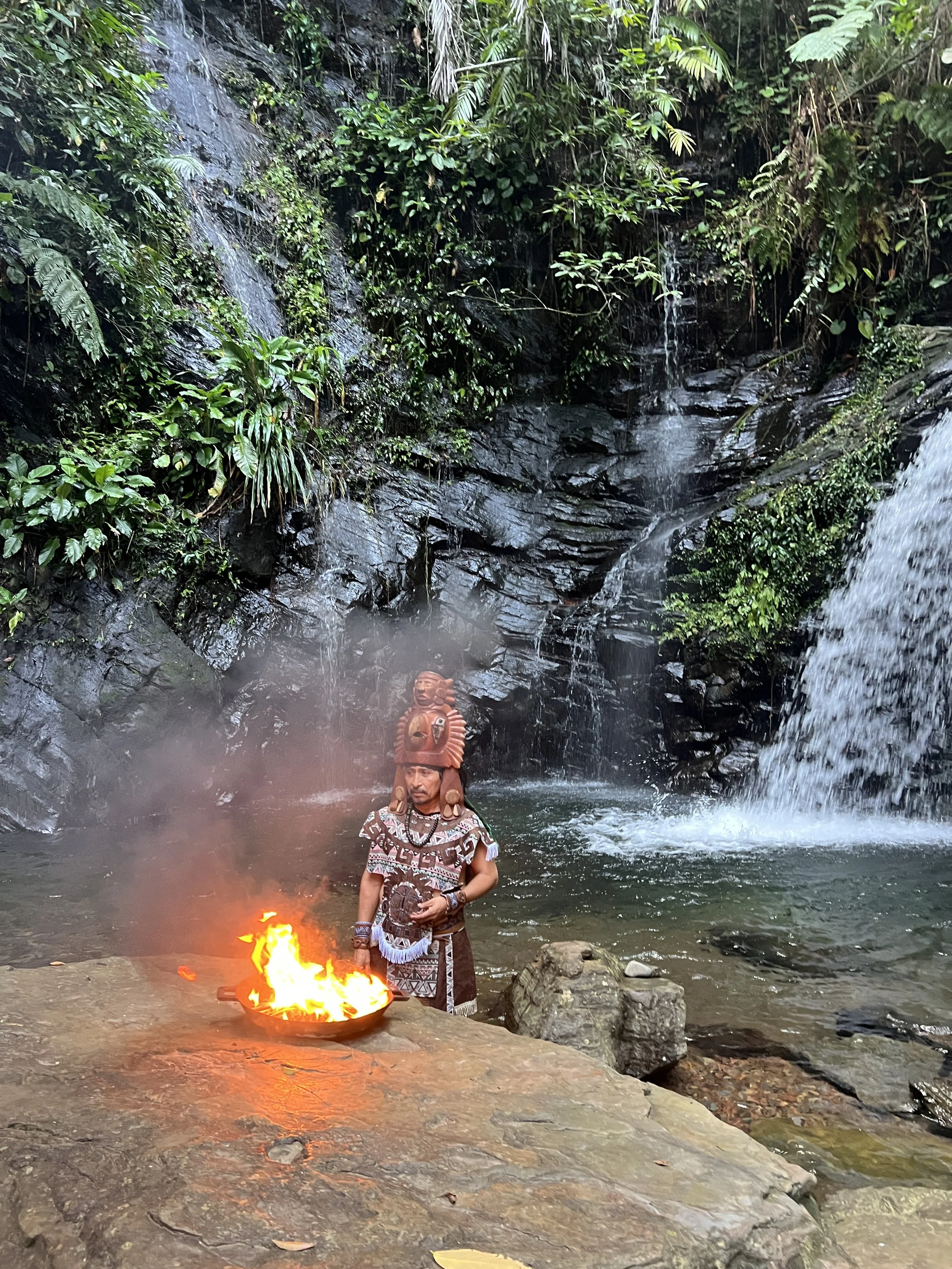 A person dressed in traditional attire performing a ritual with fire near a waterfall in a lush, tropical forest.