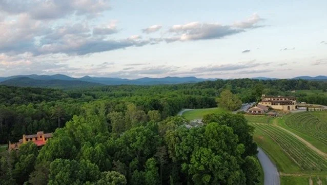Aerial view of lush green trees, a winding river, and a large building complex in a rural landscape with mountains in the distance under a partly cloudy sky.