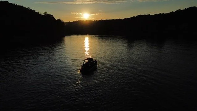 A boat on a river during sunset with the sun low on the horizon, reflecting on the water, surrounded by dark forested hills.