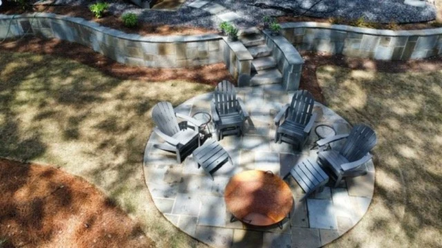 A fire pit surrounded by six gray Adirondack chairs in a backyard with a stone patio and landscaped garden.
