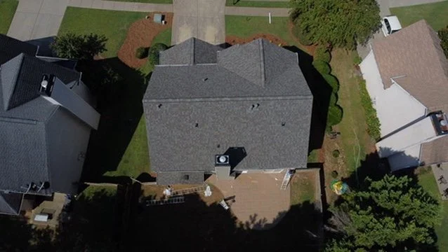 Aerial view of a house with a gray roof, surrounded by neighboring houses, a backyard with a deck, and trees.