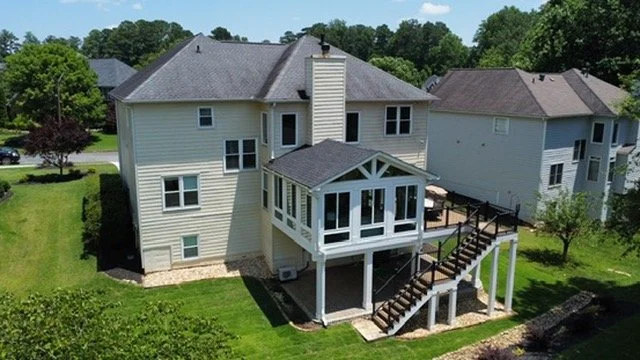 A two-story house with a gray roof and vinyl siding, featuring a screened-in porch and an outdoor staircase, in a suburban neighborhood with green lawns and trees.