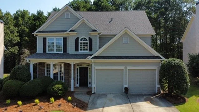 A two-story suburban house with a front lawn, driveway, and a garage, surrounded by green trees.