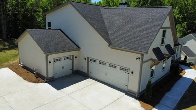 An aerial view of a modern two-story house with a three-car garage, gray roof, and white exterior, surrounded by a concrete driveway and a landscaped yard.