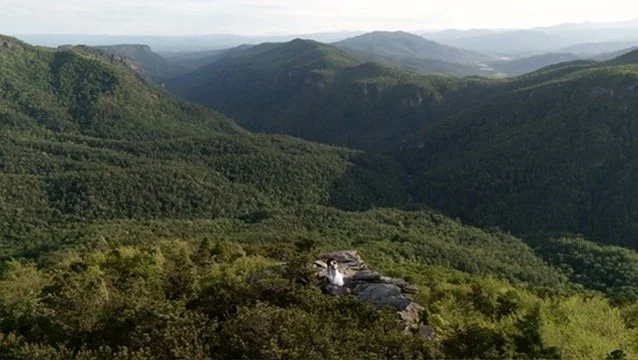View of lush green mountains and valleys with a person sitting on rocks near the edge of a cliff, overlooking the forested landscape.