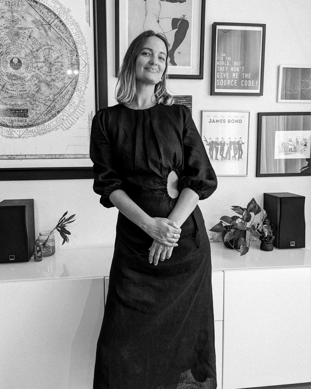 A woman in a black dress standing in front of a wall decorated with framed artwork, posters, and a large astrological chart, with two speakers and potted plants on a white shelf.