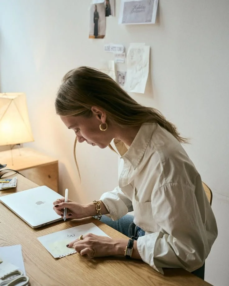 A woman sitting at a desk writing on a piece of paper with an Apple MacBook beside her. There is a lamp on her left and pictures and notes on the wall behind her.
