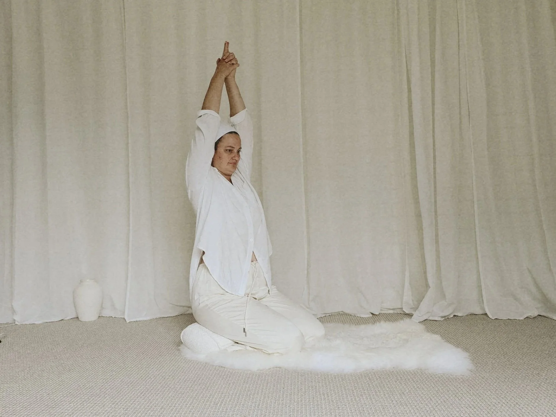 A woman dressed in white, kneeling on a soft rug with her hands raised above her head in a prayer position, in front of light-colored curtains.