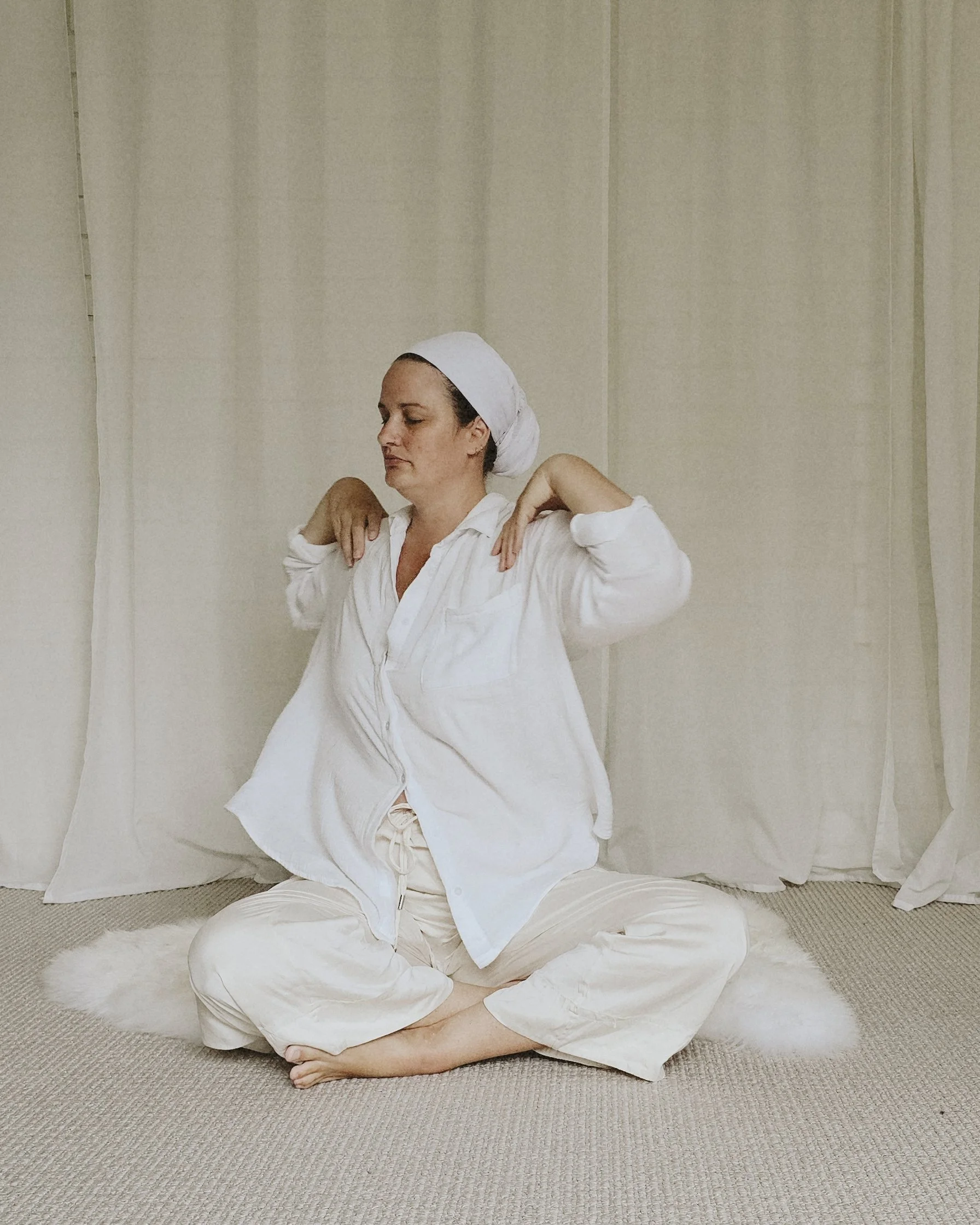 A woman sitting cross-legged on a plush white rug, wearing white pajamas and a head scarf, meditating with her hands resting on her shoulders in front of a neutral-colored curtain background.