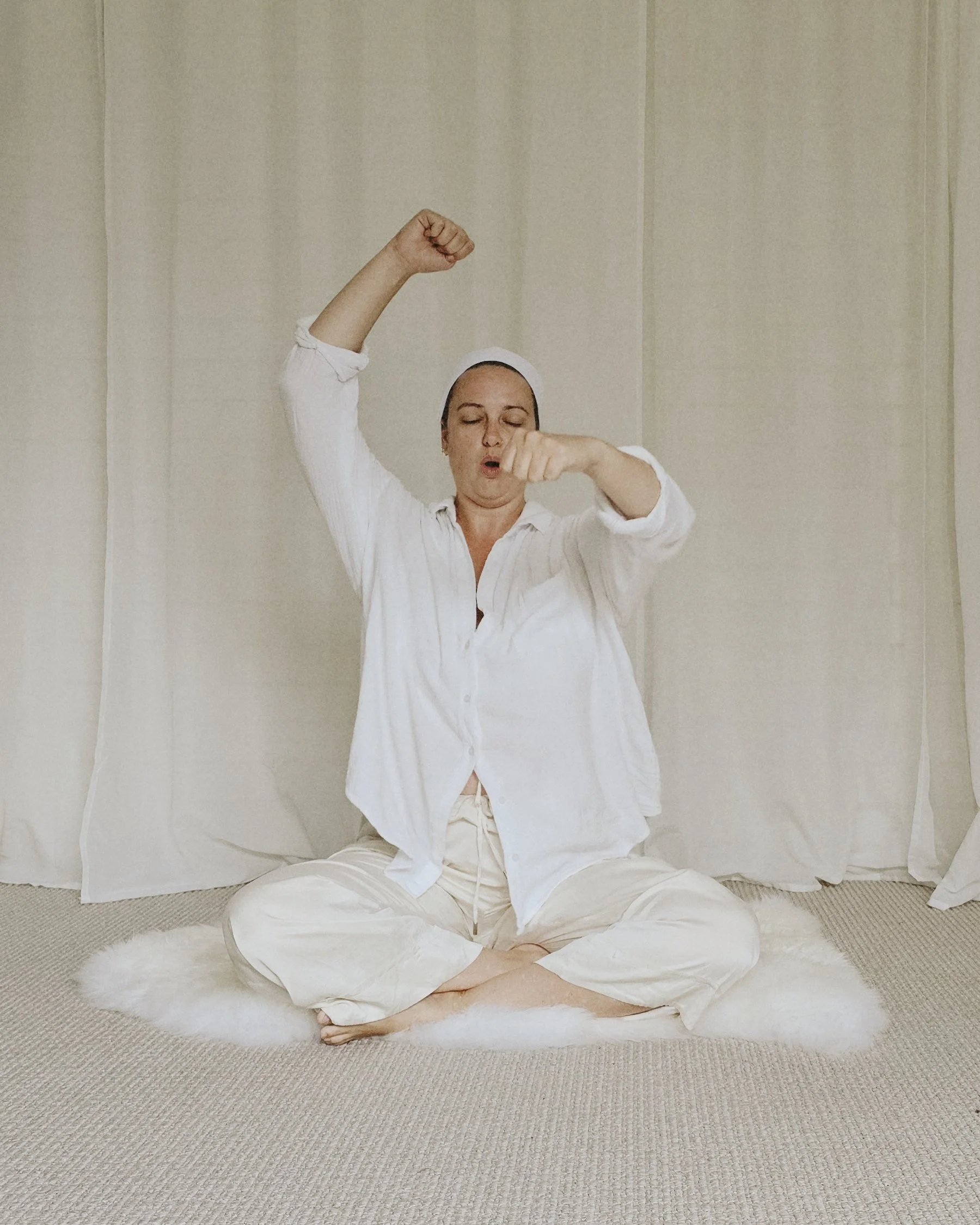 Woman dressed in white loungewear sitting cross-legged on a white fur rug, raising one arm and making a fist, with eyes closed and an expressive face, in front of cream-colored curtains.