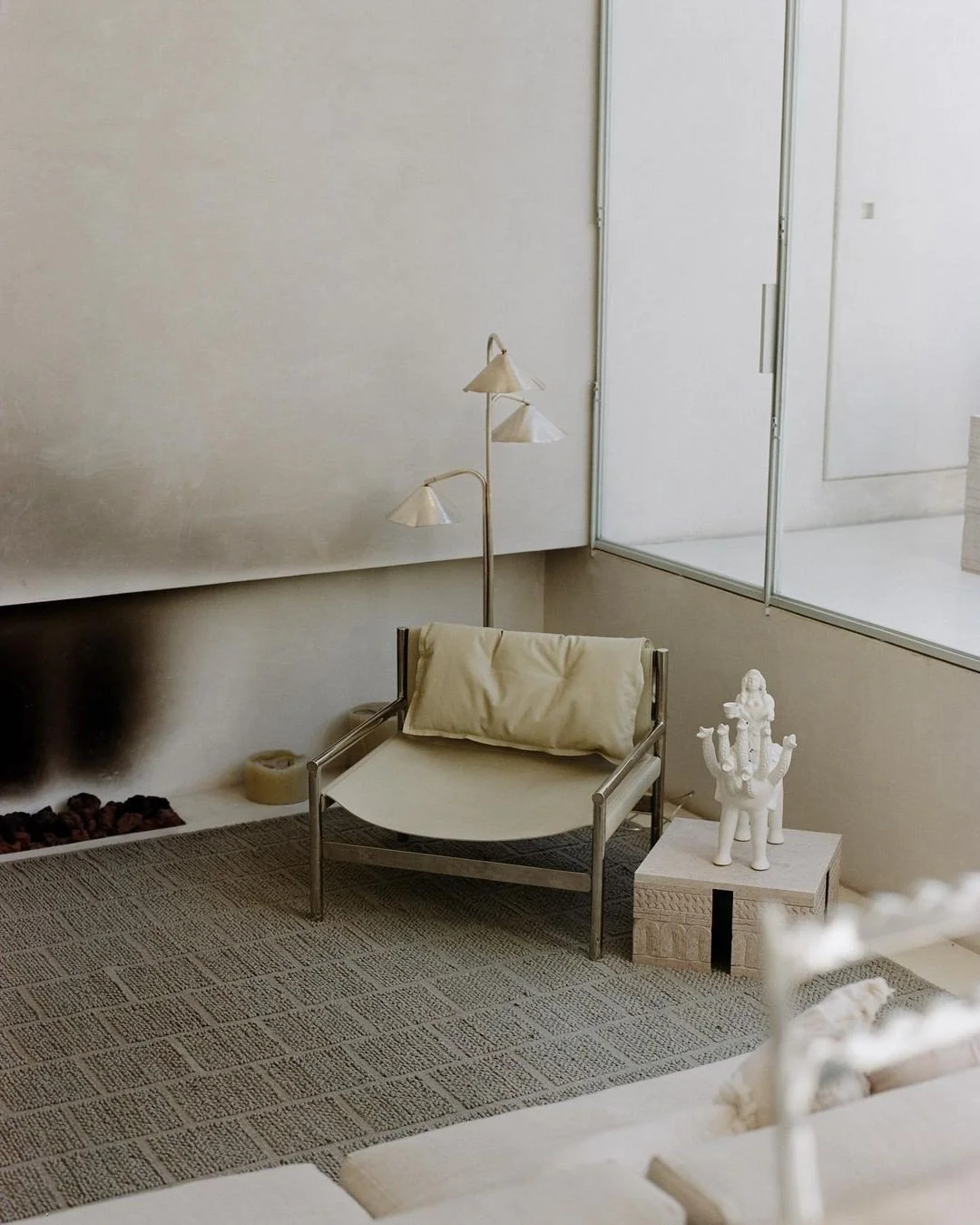 A contemporary interior space with a metal-framed chair with cream cushion and a matching pillow, a side table with a decorative white sculpture resembling a multi-armed figure, a textured rug, a floor lamp with three cone-shaped shades, and a large glass window or wall showing an adjacent room with white walls.