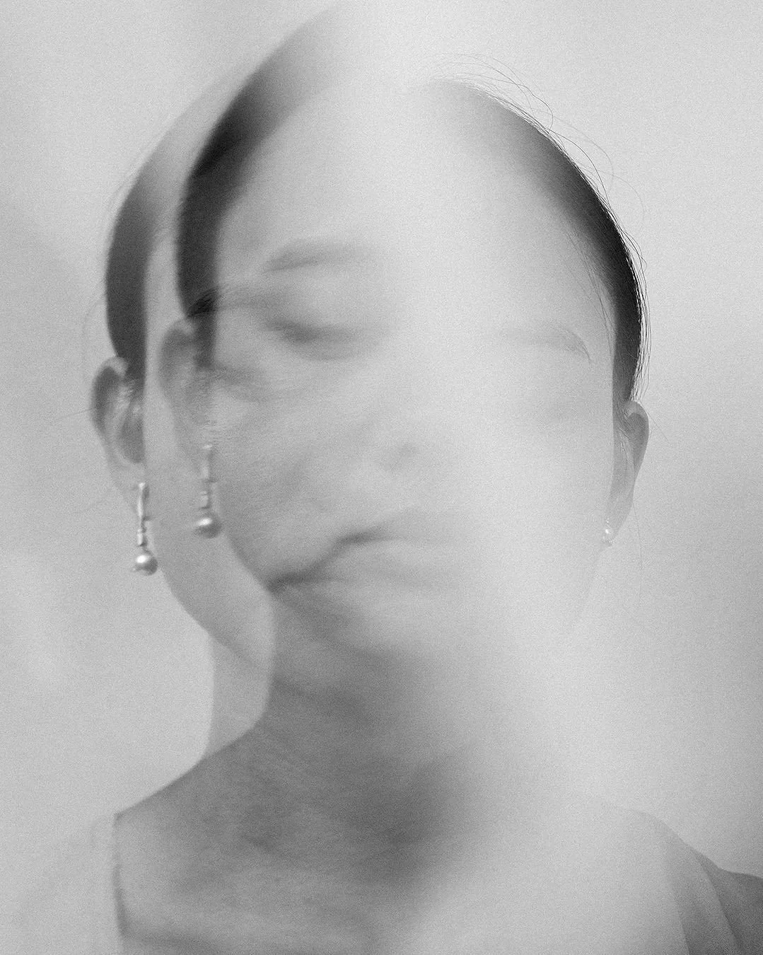 A black and white photo of a woman with earrings and her eyes closed, with a blurred, double exposure effect.