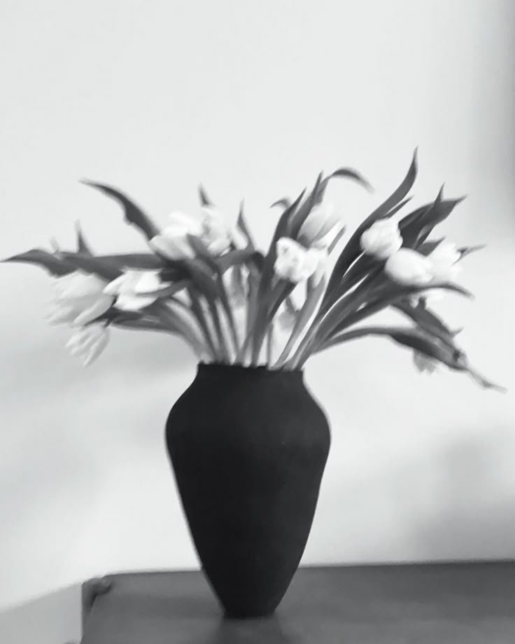 A black vase holding a bouquet of white flowers, placed on a wooden surface against a plain wall background.