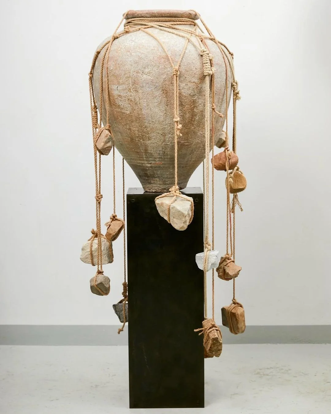 Ancient clay pot wrapped with rope and rocks, displayed on a black pedestal in a gallery setting.
