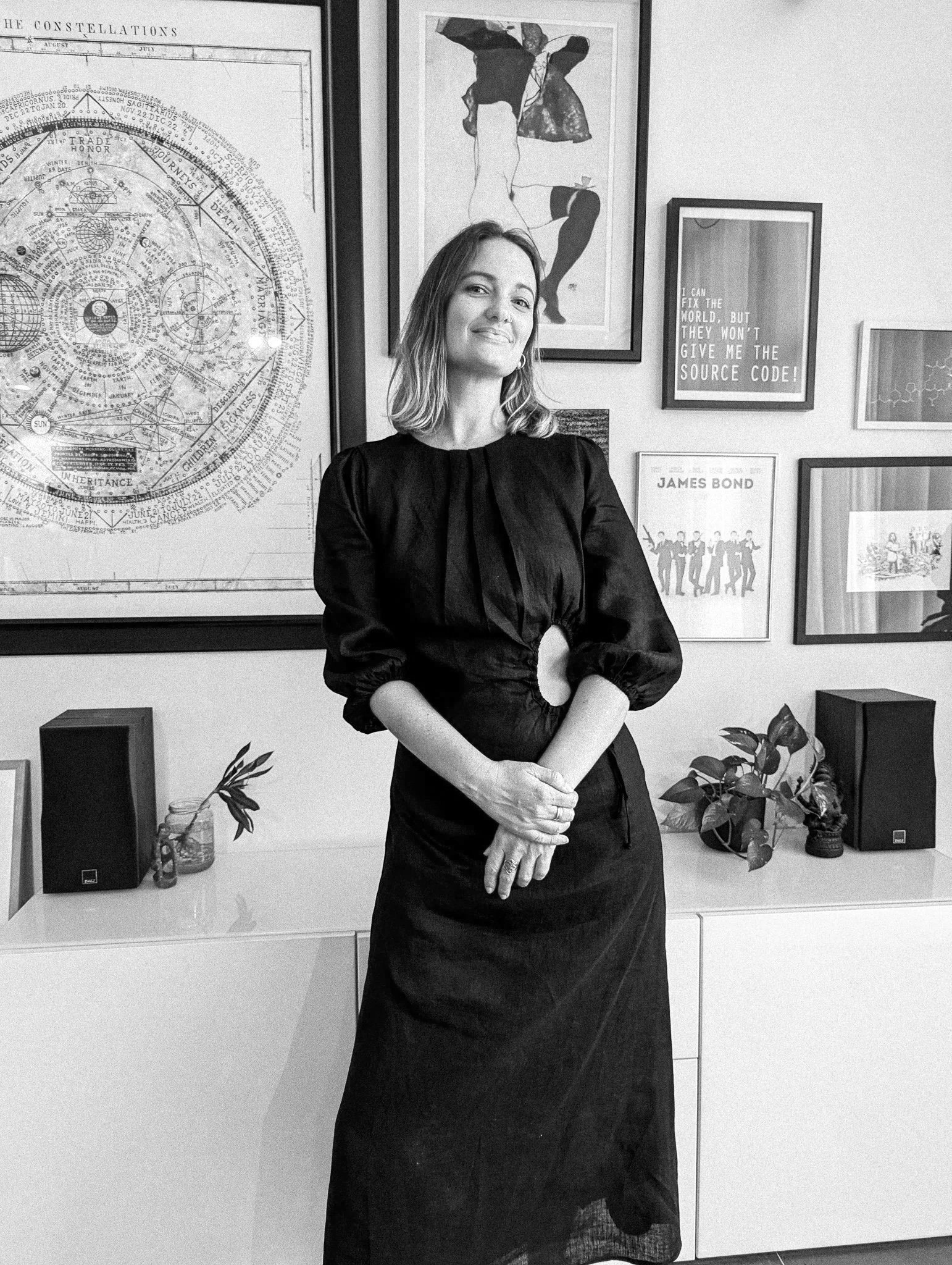 A woman standing in front of a gallery wall decorated with framed posters and artwork, including star charts, a nude figure, and a quote. She is wearing a long black dress with arched sleeves and has shoulder-length hair, smiling softly with her hands clasped.