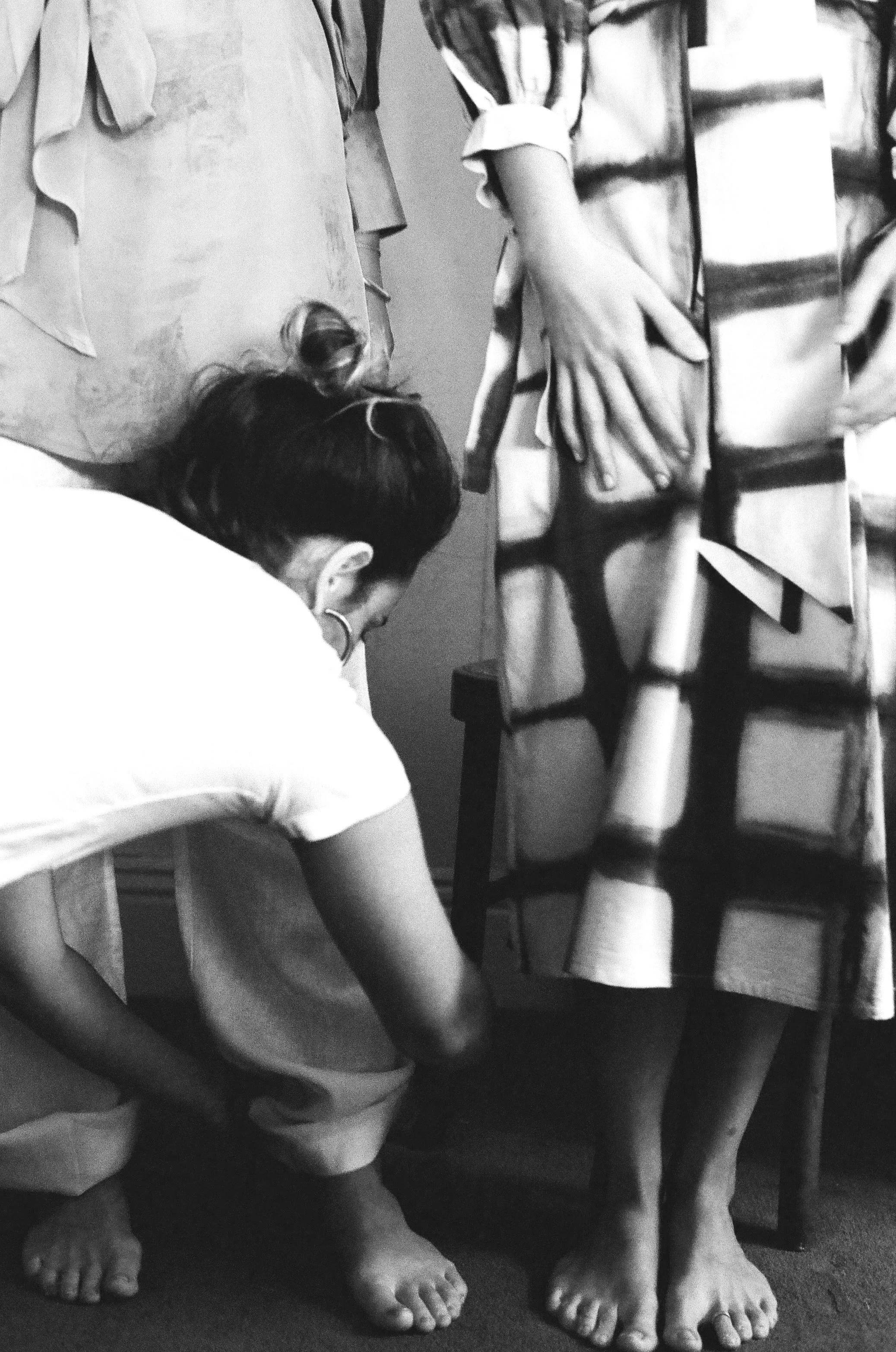 A woman crouching down on the floor, with her head bowed, next to another woman standing with her hands on her hips, both in a room filled with clothing and fabric items.