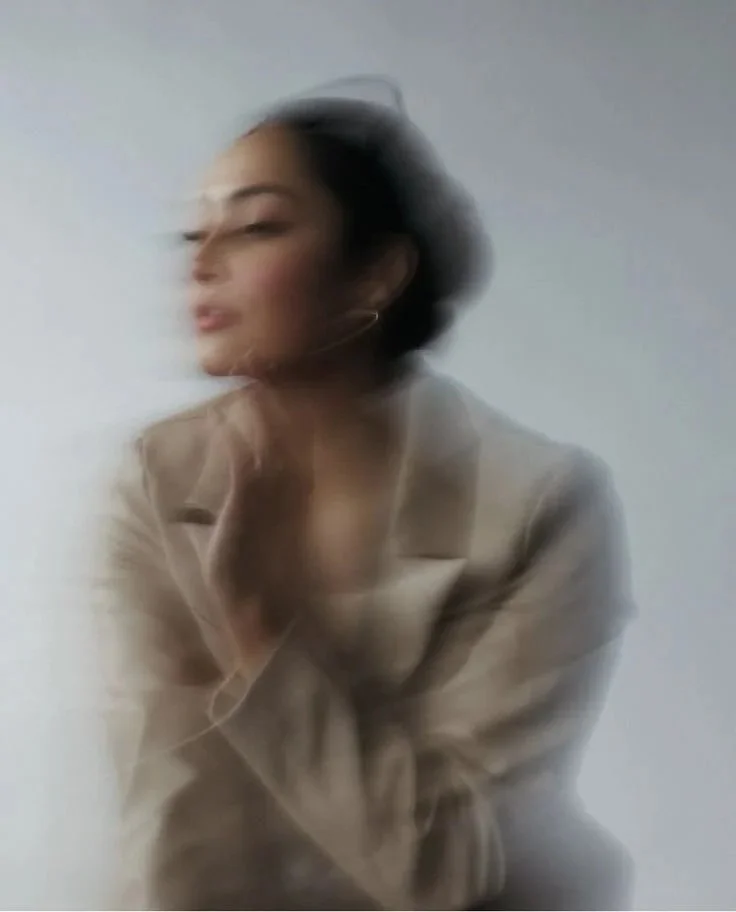 Blurred image of a woman with short black hair wearing a beige blazer, sitting against a plain light background.