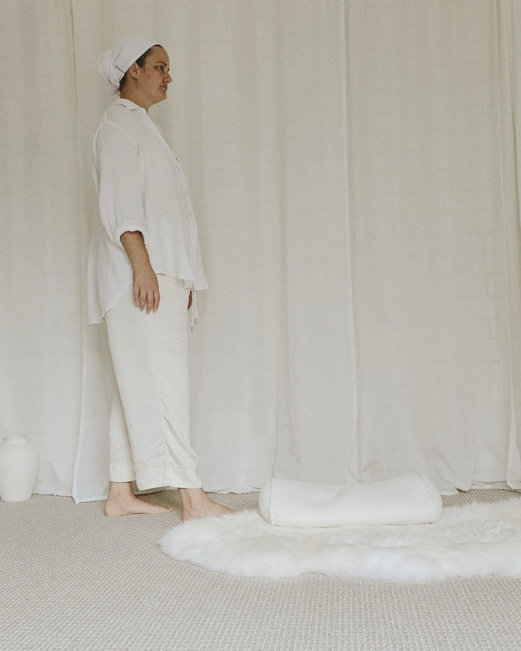 Woman dressed in white, standing barefoot on a white rug, against a cream-colored curtain background.