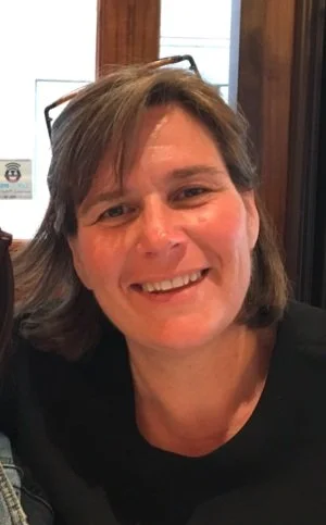 A smiling woman with short brown hair, wearing glasses on her head, sitting indoors in front of a window and wooden paneling.