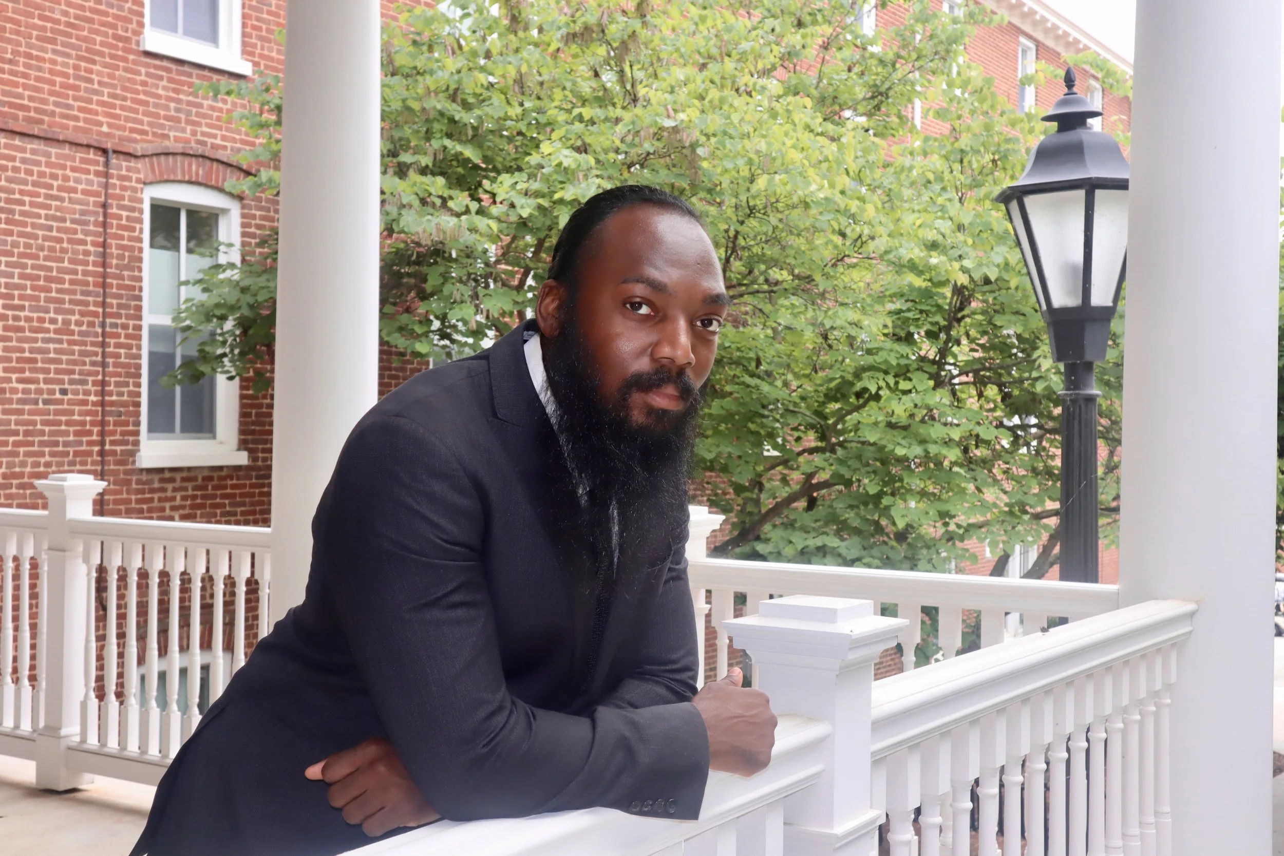 A bearded man in a black suit leaning on a white porch railing, with a brick building, green trees, and a street lamp in the background.