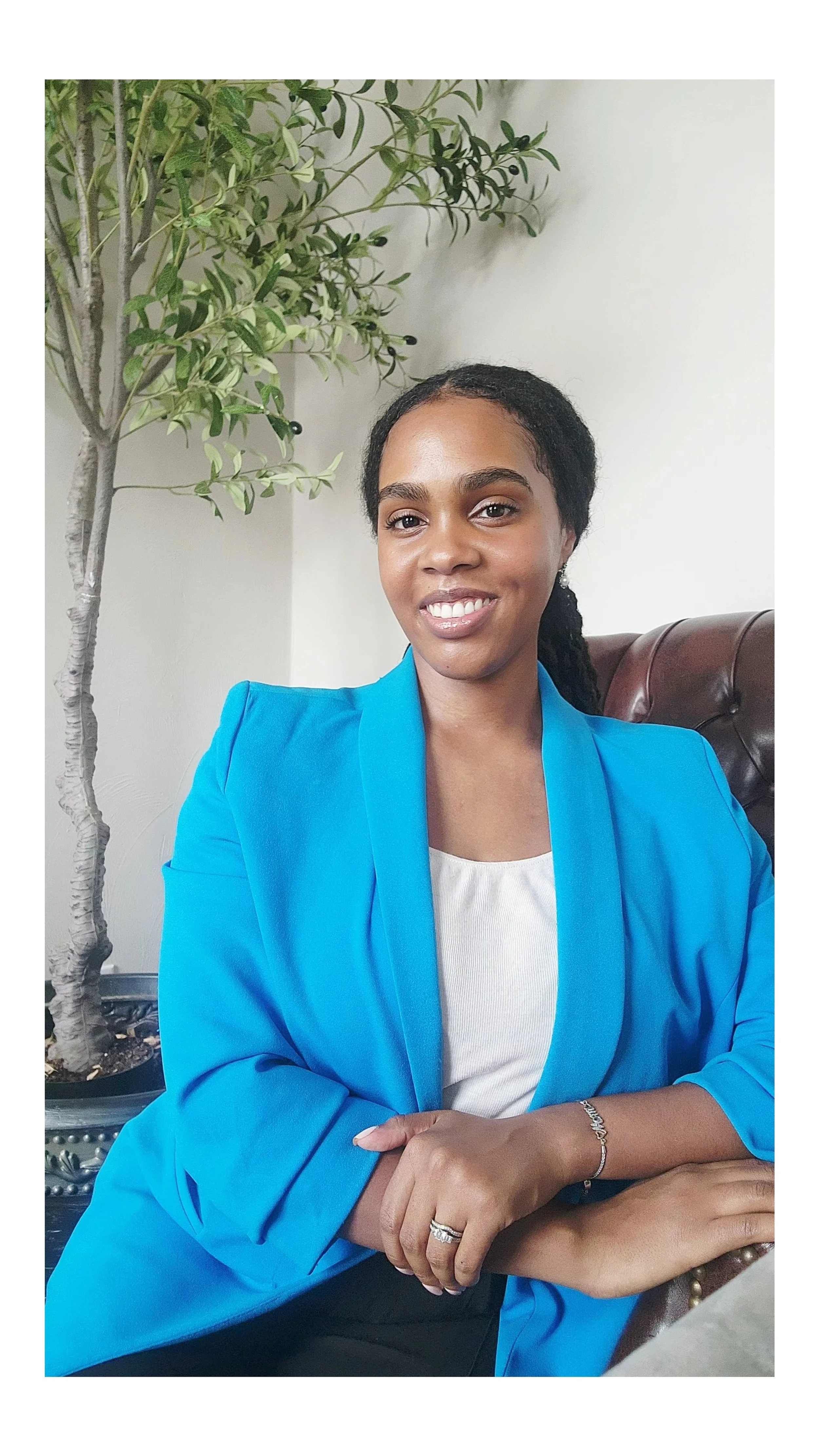 A woman with dark curly hair smiling, wearing a blue blazer and a white top, sitting on a brown leather chair with a green potted plant beside her.