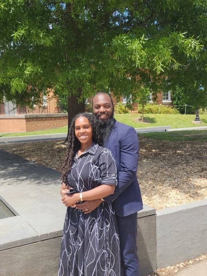 A smiling man and woman standing outdoors under a large green tree, with the man hugging the woman from behind. The woman is wearing a black dress with white swirly lines, and the man is dressed in a navy blue suit. They are in a sunny area with a brick building in the background.