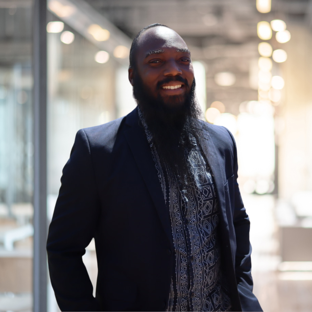 A man with a beard and dark skin, wearing a dark blazer over a patterned shirt, smiling in a modern indoor space with blurred lights in the background.