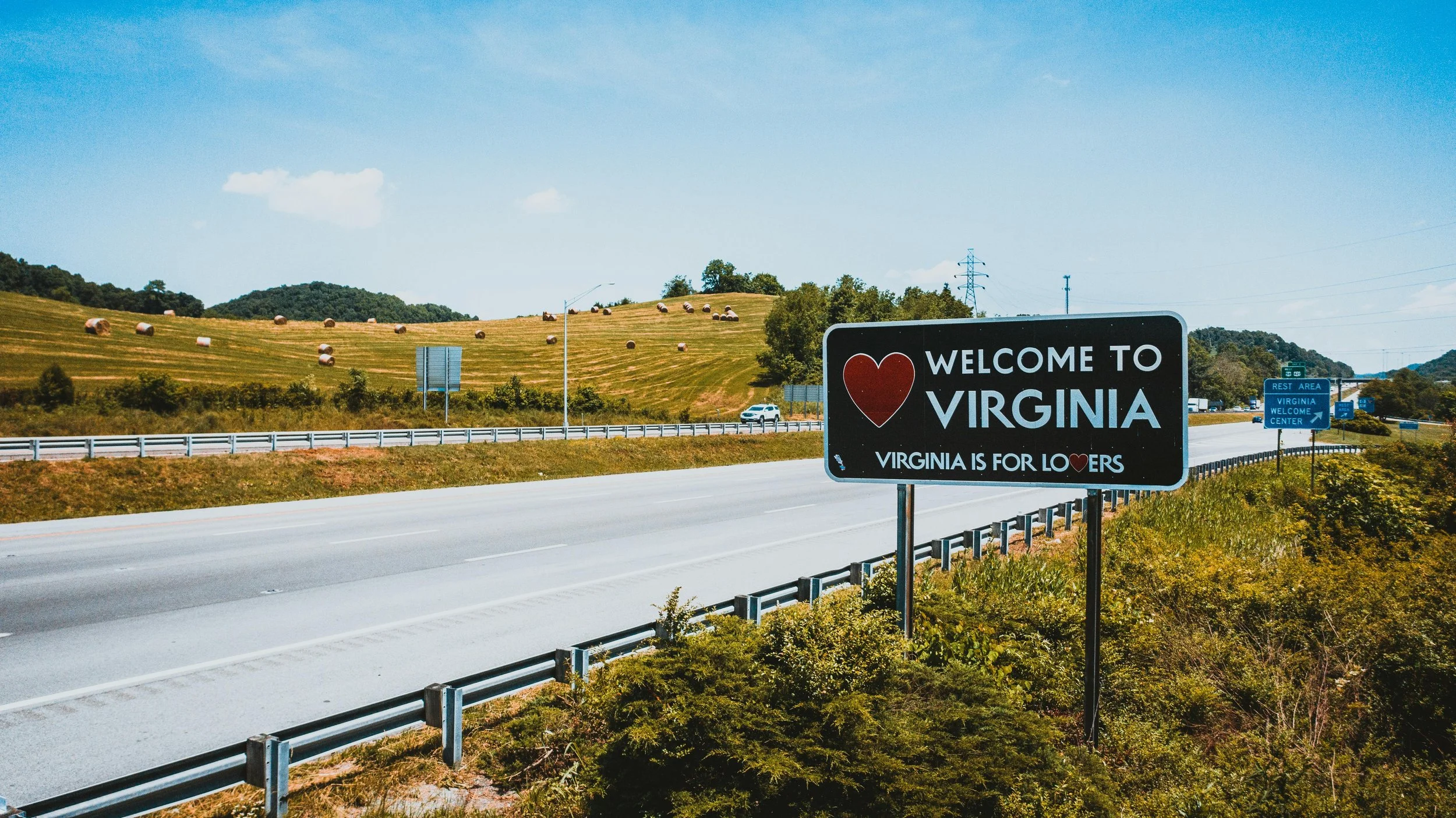 A highway leading into Virginia with a sign that reads "Welcome to Virginia, Virginia is for Lovers". The sign features a red heart and is in the foreground. There are rolling green hills with hay bales in the background under a blue sky.