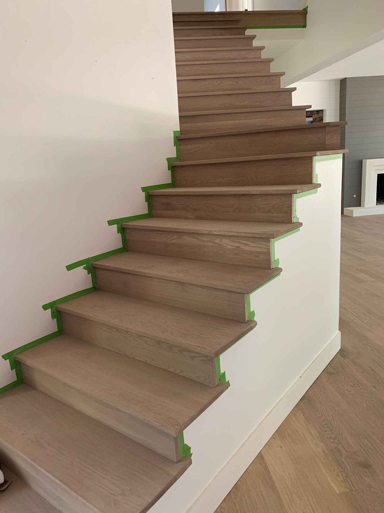 Wooden staircase with light brown steps, partially taped with green painter's tape on the edges, in a modern home interior.