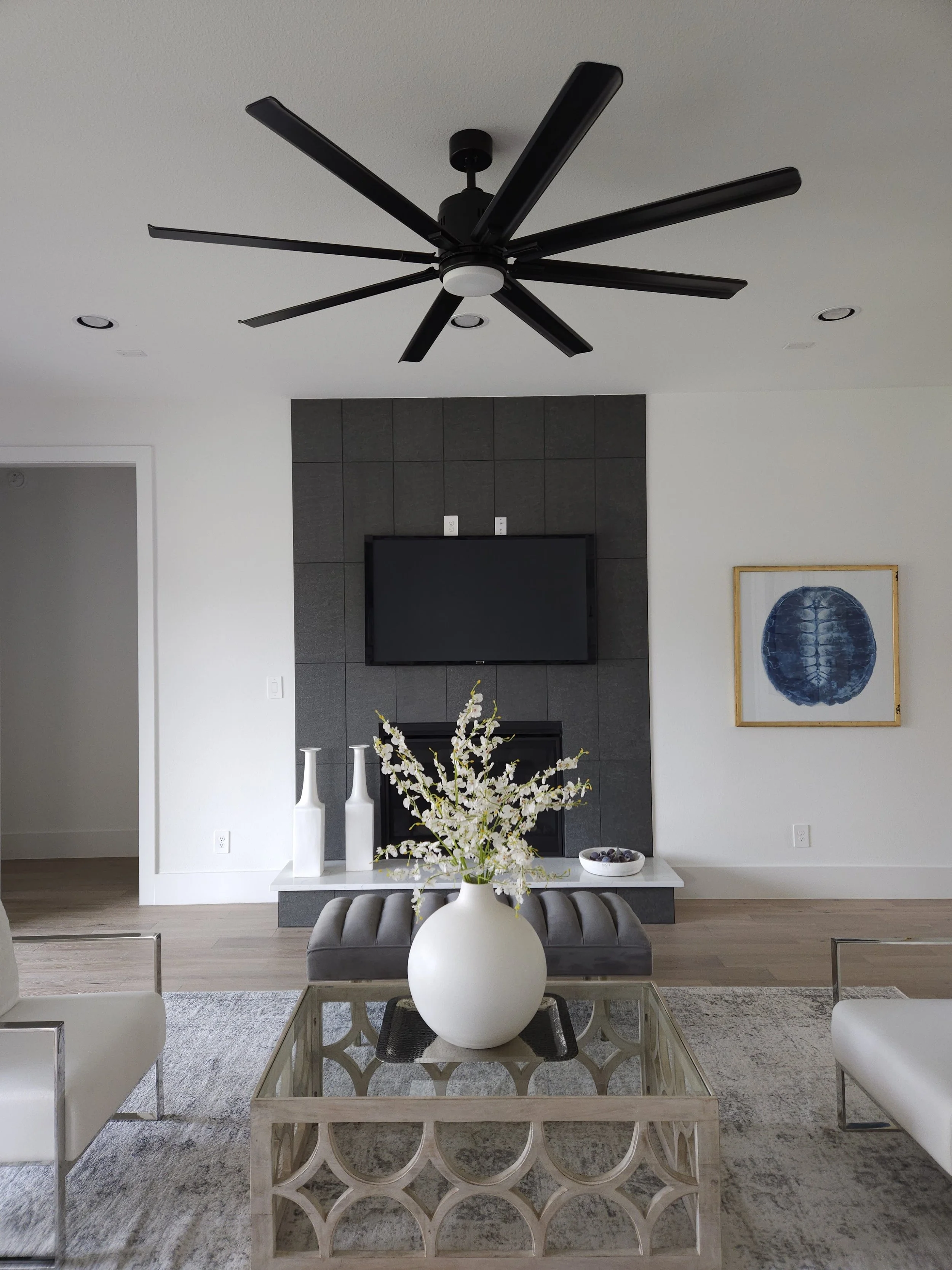 Modern living room with a black ceiling fan, white walls, grey tile fireplace with a television, white and blue art, and a glass coffee table with white vases and a floral arrangement.