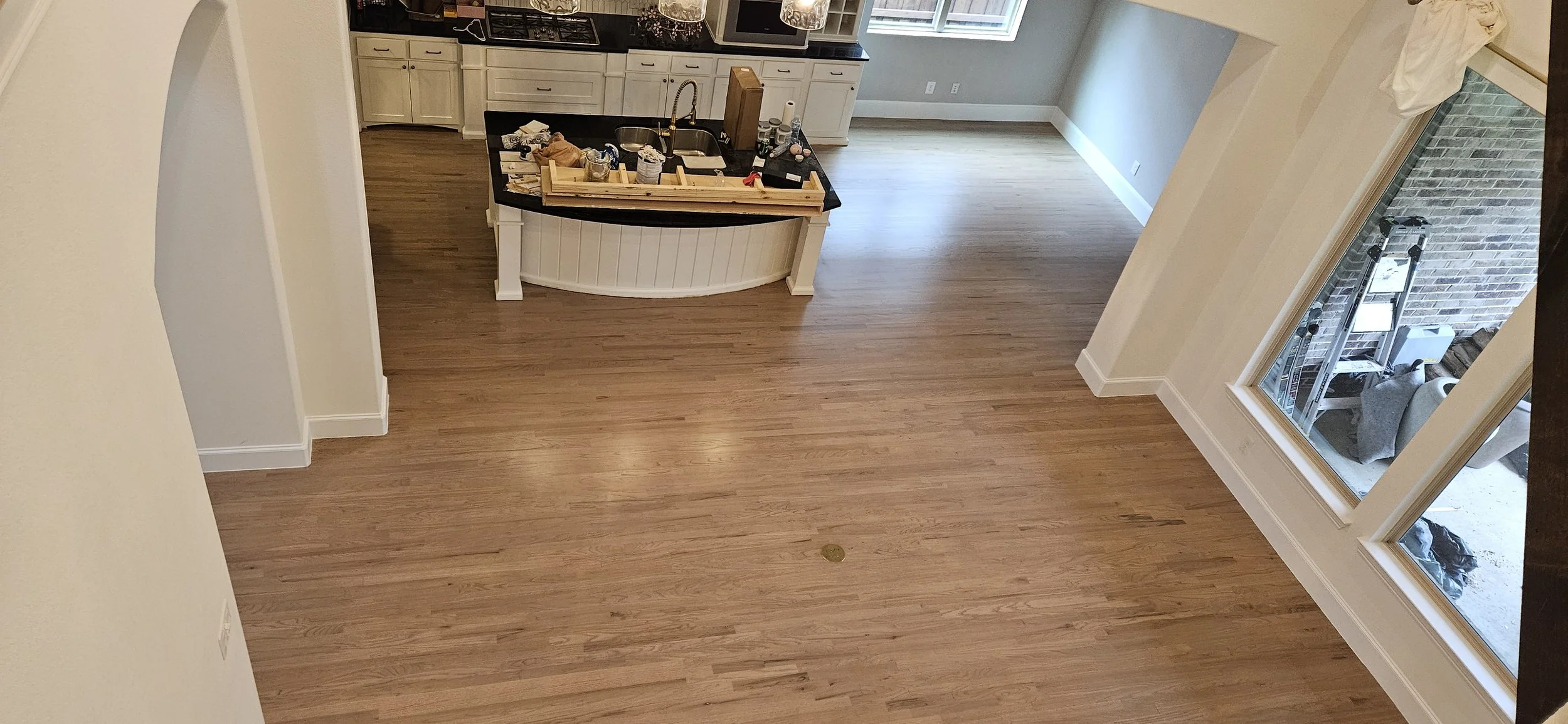 Empty room with hardwood floors, kitchen in the background with cabinets, and a large window looking into an outdoor area with construction tools and a ladder.