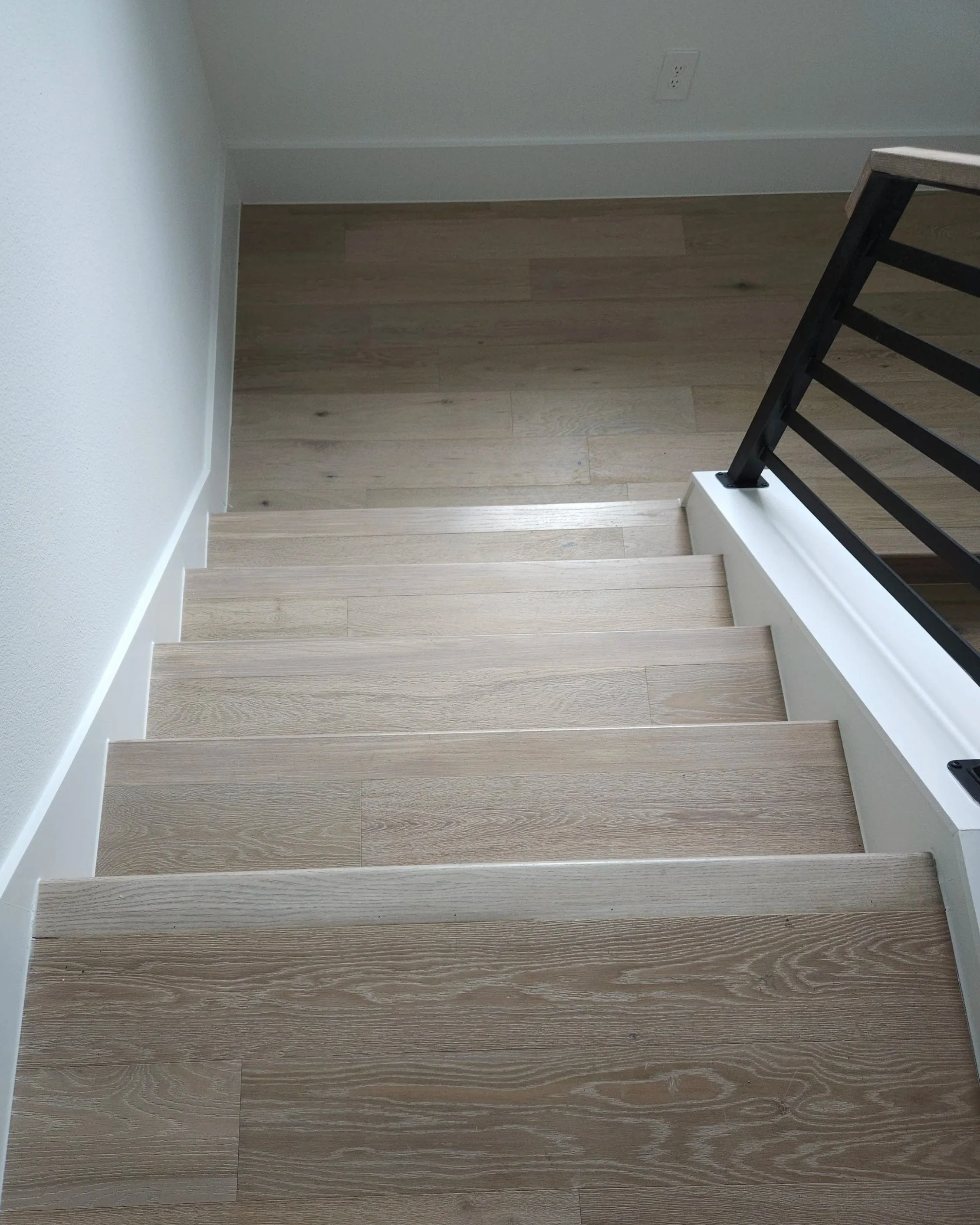 Top view of wooden staircase with black railing, white baseboards, and hardwood flooring at the bottom, with a person's feet visible at the top