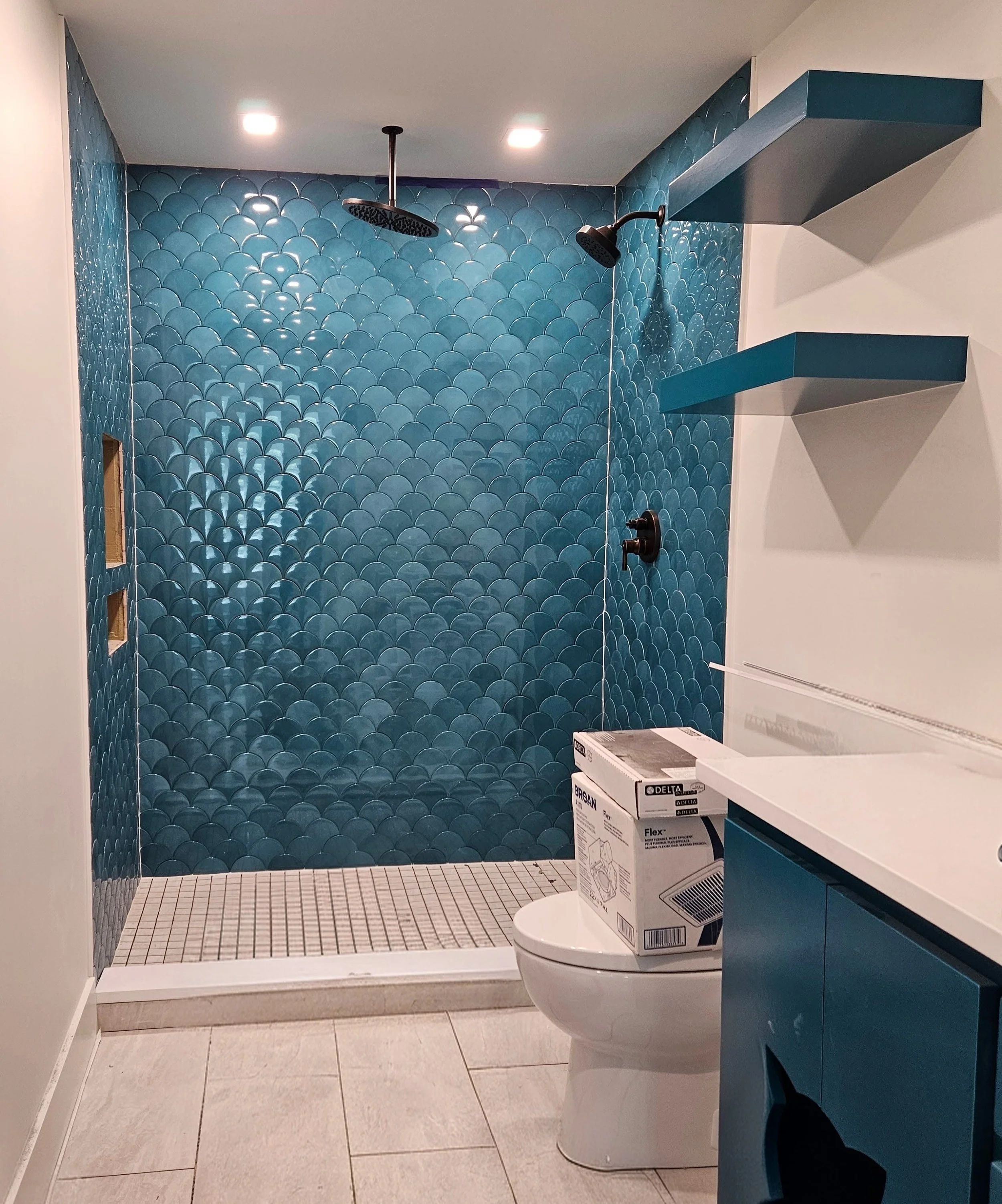 Bathroom with a walk-in shower area featuring blue scalloped tiles, a rainfall showerhead, and a wall-mounted showerhead. There's a white toilet with a box on top, a blue cabinet, and floating blue shelves. The floor has light-colored tiles.