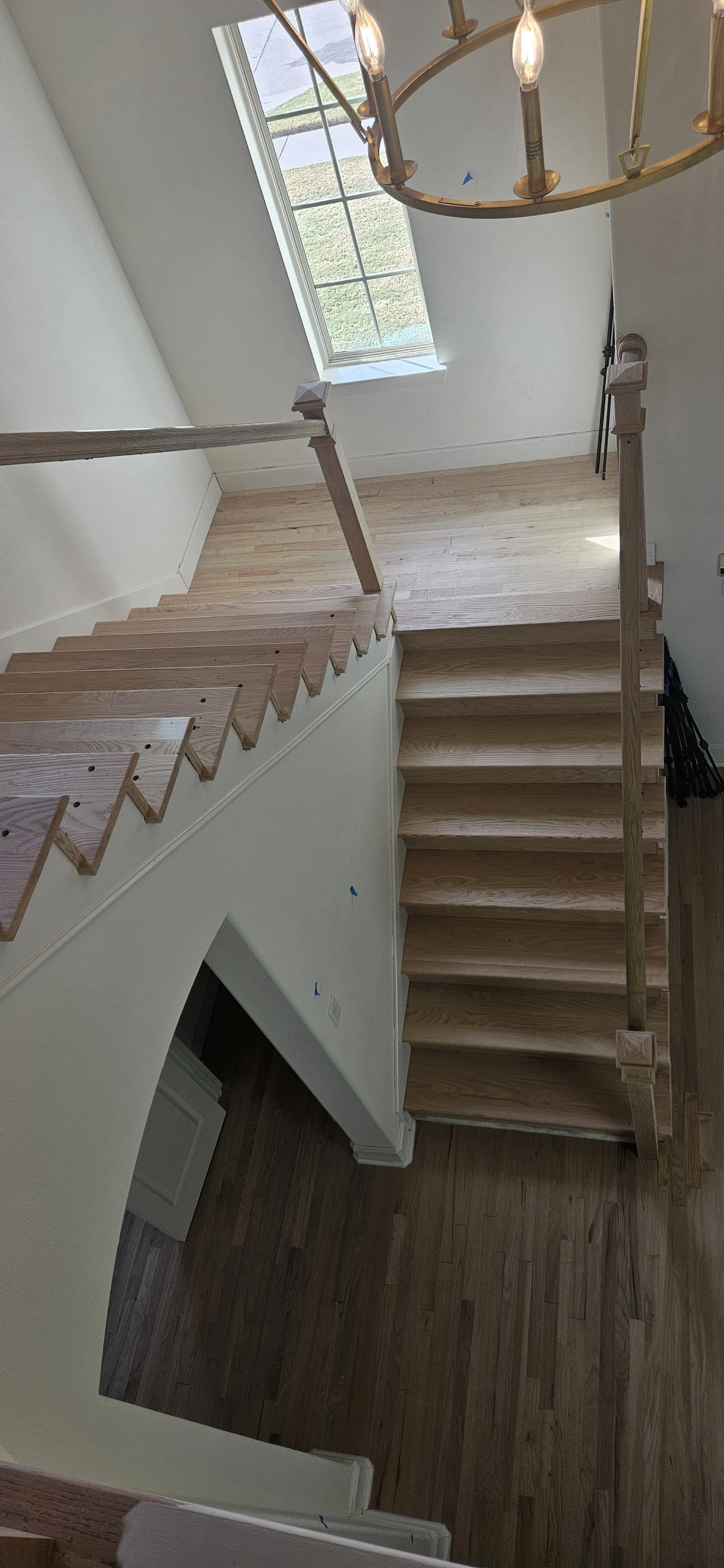 View of a staircase with unfinished wooden steps and matching handrails, leading down from a second floor to a lower level, seen from above, with a large window at the top letting in natural light, a chandelier hanging from the ceiling, and wooden fl