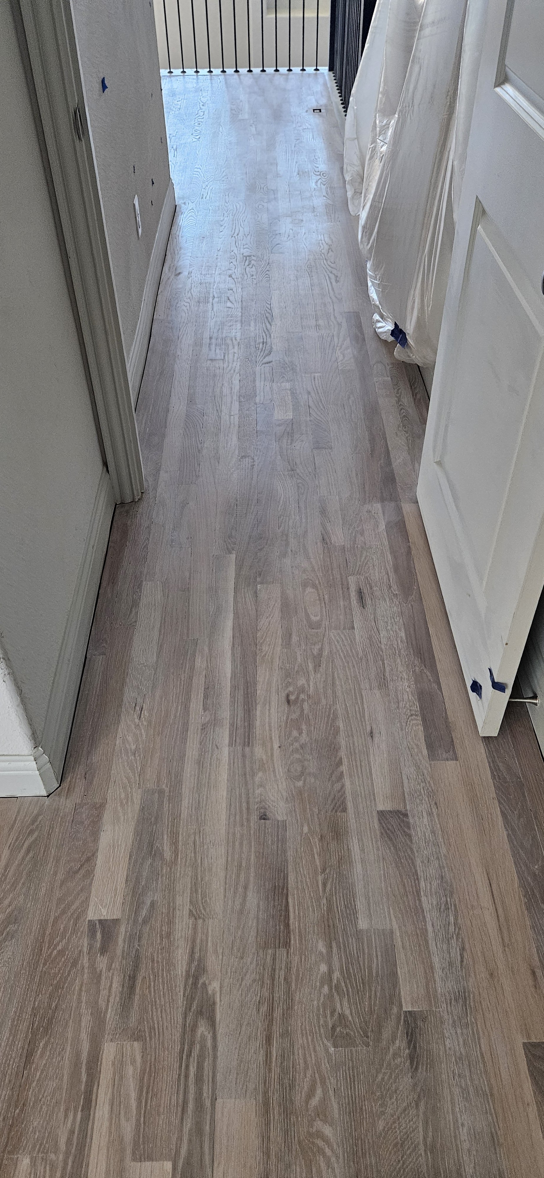 Newly installed light-colored hardwood floor in a hallway near a balcony with a black railing. Part of a door with protective cover on the right side of the image.