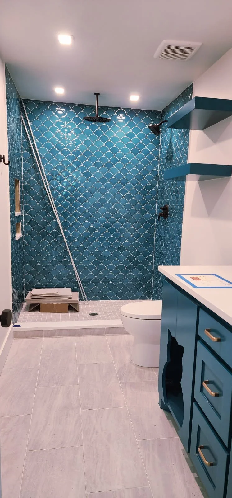 Bathroom with blue and white scalloped tile shower wall, ceiling-mounted showerhead, toilet, blue vanity with gold handles, and open shelves with blue accents.