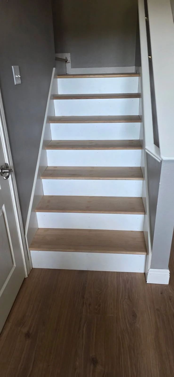 A staircase with light wood steps and white risers, located between gray walls, with a small section of a white door visible to the left.