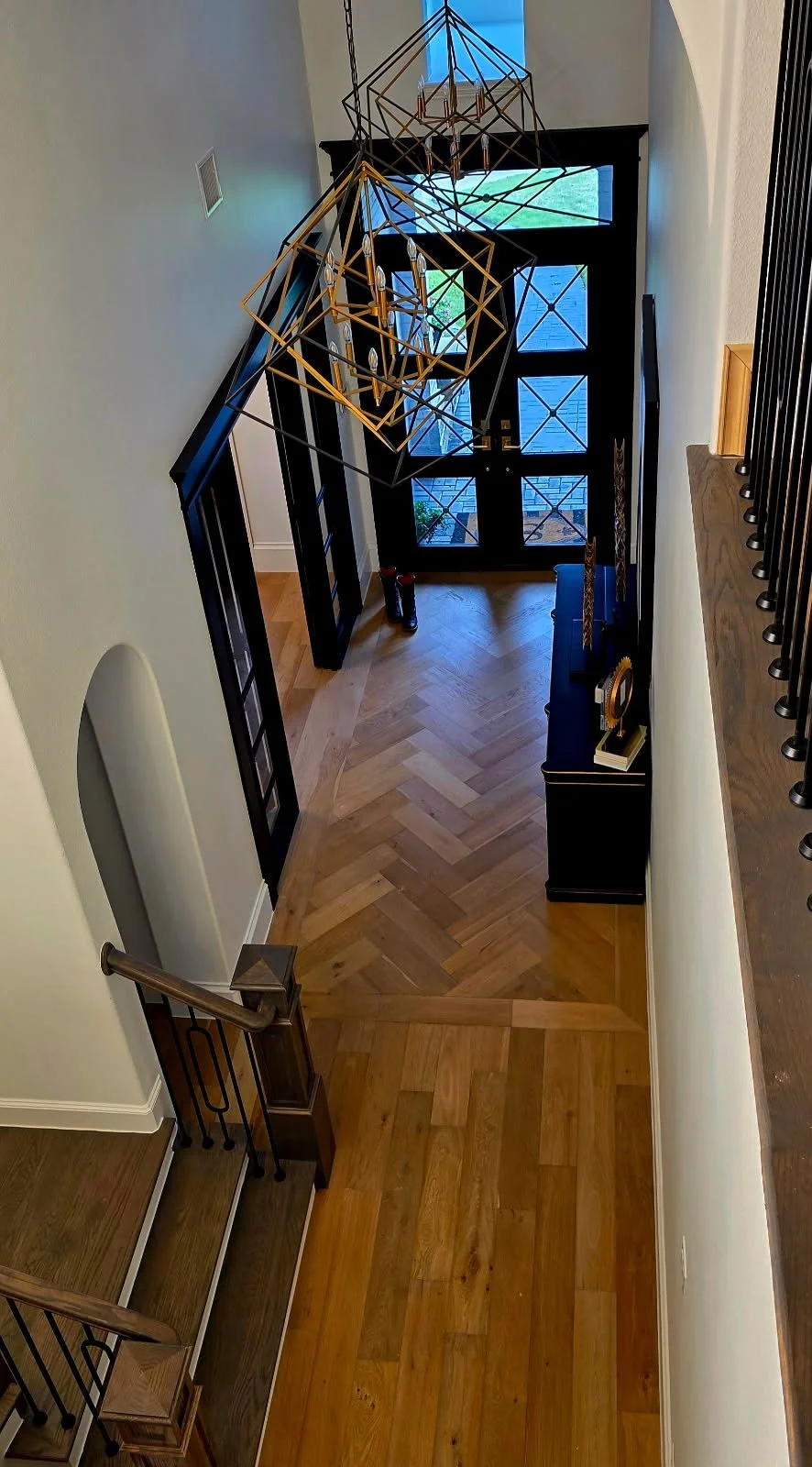 View of a home's entryway with a black front door and glass panels, hardwood flooring in a herringbone pattern, and modern gold geometric chandelier lighting fixtures hanging from the ceiling.