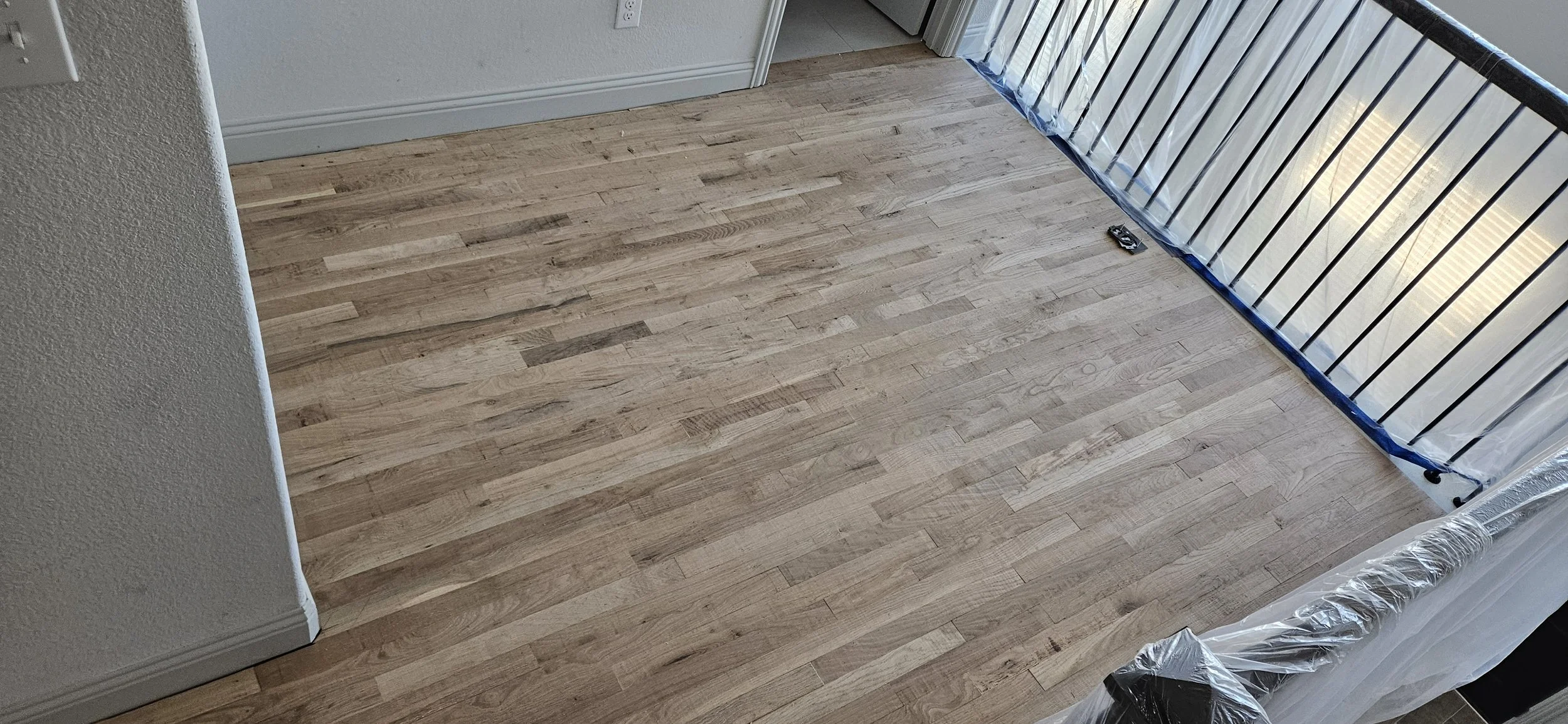 Newly installed light-colored hardwood flooring in a corner of a room, with a balcony railing on one side and a wall with a door on the other, and some plastic wrapping along the edge.