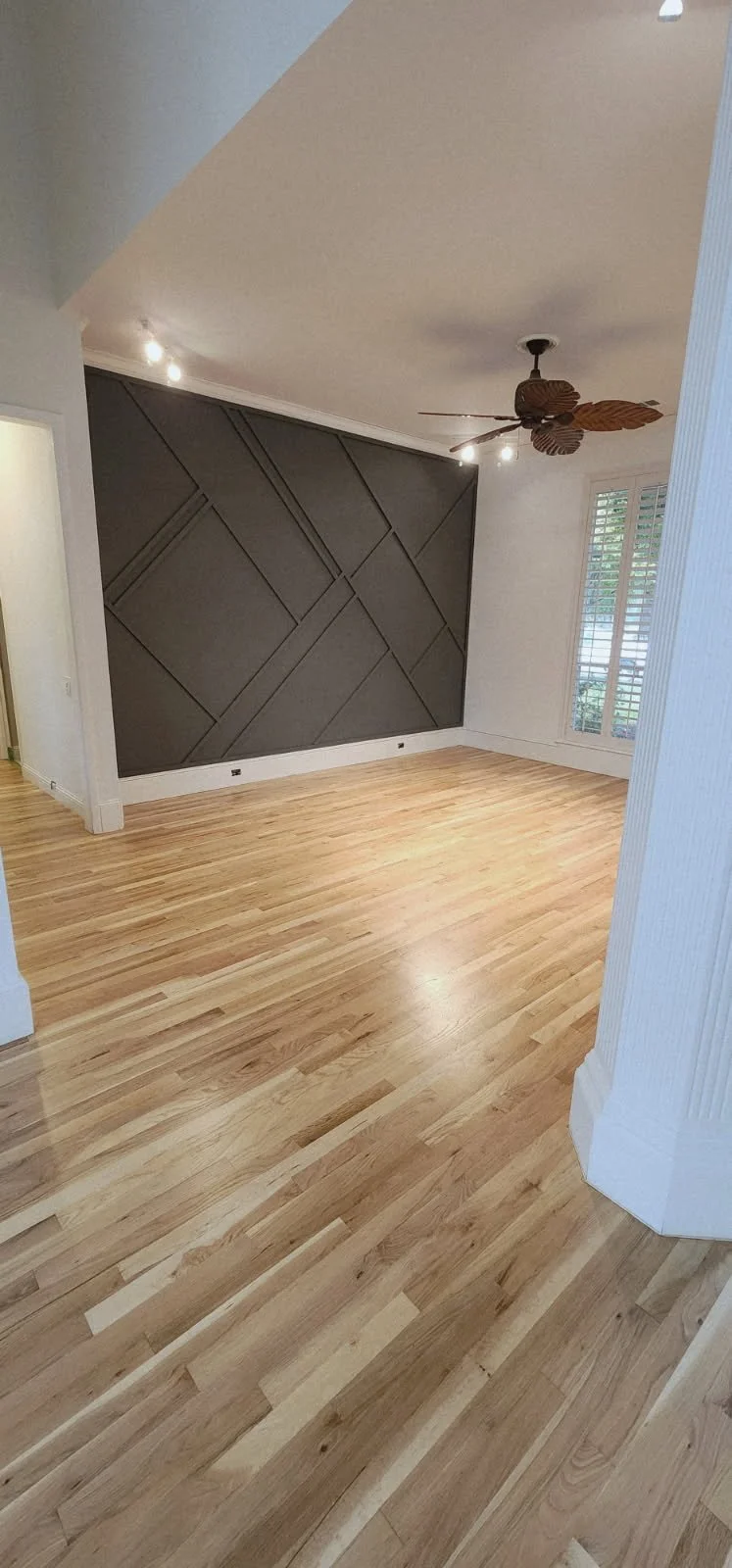 Empty room with wooden floors, a dark accent wall with geometric paneling, a ceiling fan with leaf-shaped blades, and a large window with white shutters.