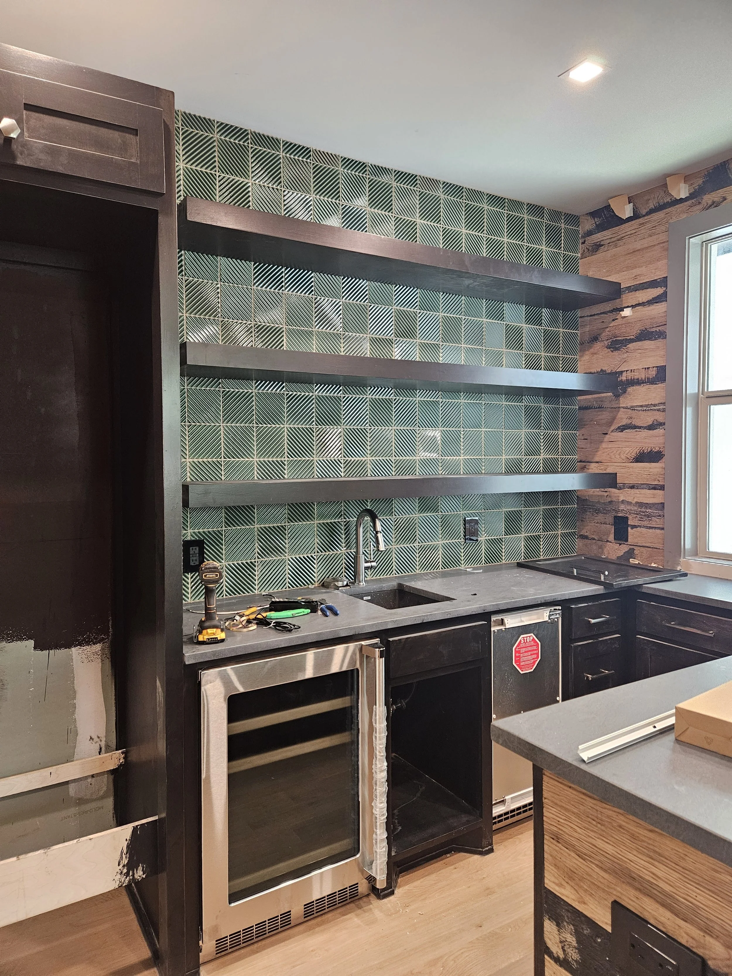 Kitchen with black cabinets, a granite countertop, green patterned tile backsplash, and open wooden shelves, under construction.