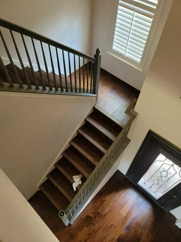 Interior view of a staircase and hallway with wooden floors, a window with white shutters, and a black door with a glass panel.