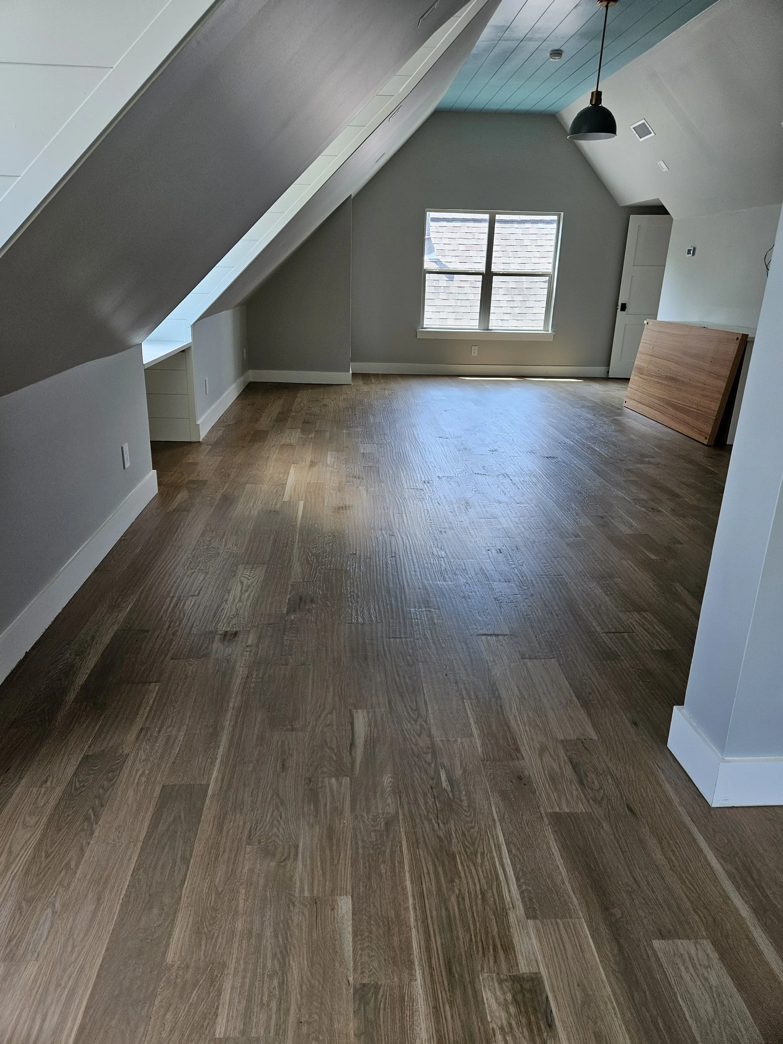 Empty attic room with sloped ceiling, wooden flooring, window, door, and a hanging black pendant light.