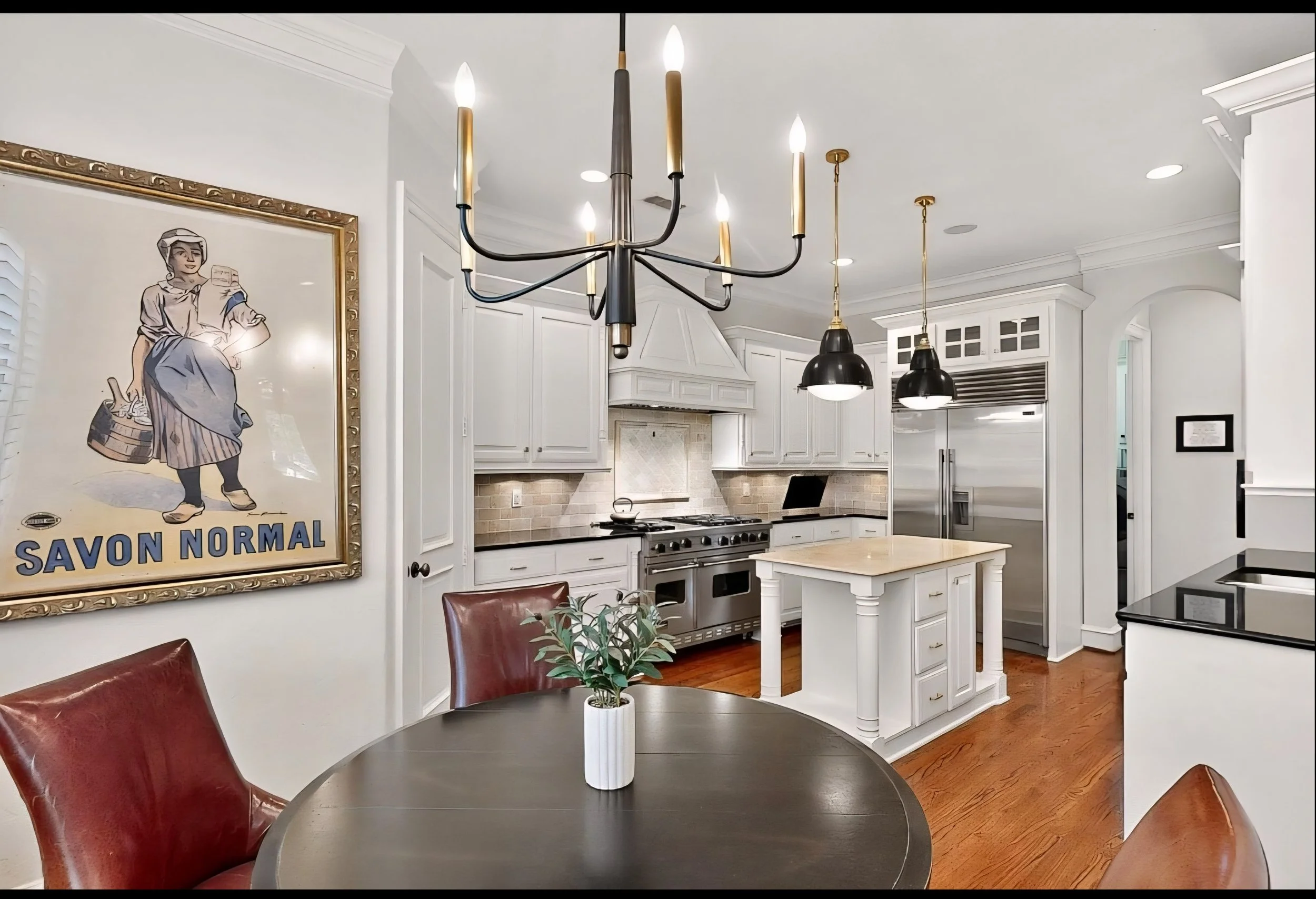 A modern kitchen with white cabinets, stainless steel appliances, a small kitchen island, a round dining table with brown leather chairs, hanging pendant lights, and a framed vintage poster on the wall.