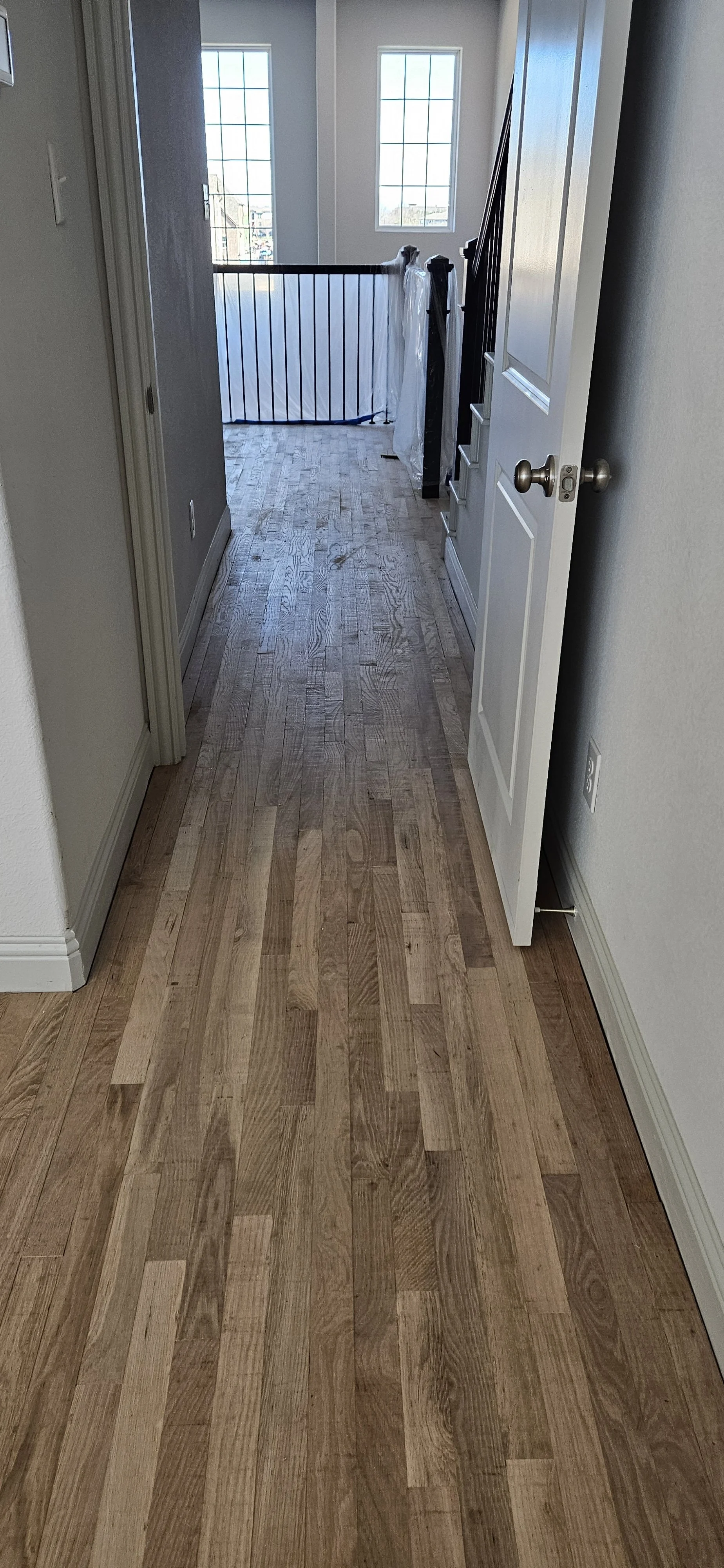 View of a hallway with wooden flooring leading to an open space with large windows, a staircase with a partially covered newel post, and a white door on the right side.
