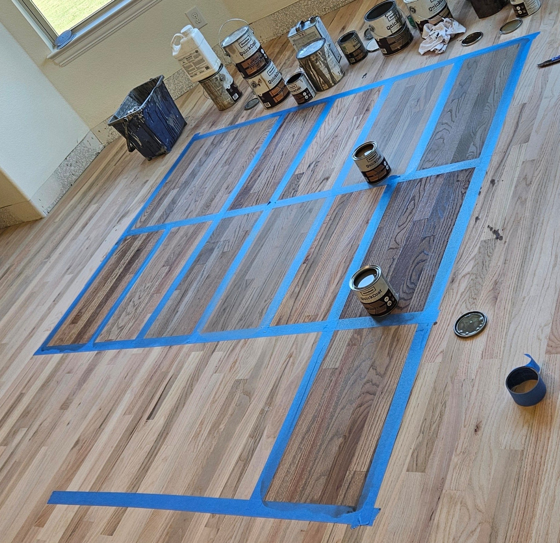 A hardwood floor with blue painter's tape creating a grid pattern, with several cans of paint and painting supplies around the area.