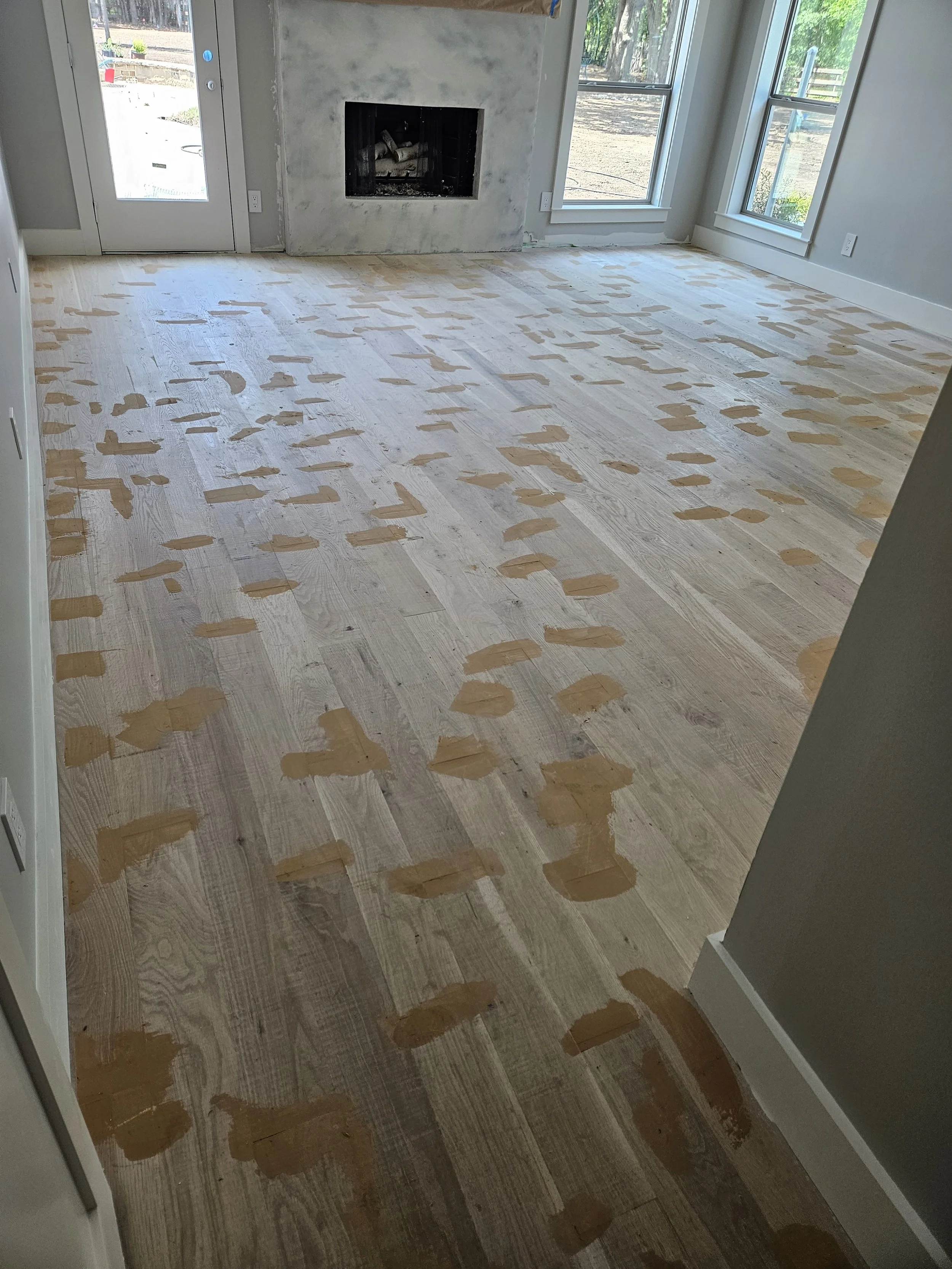 Freshly sanded hardwood floor in a living room with a fireplace, large windows, and a glass door leading outside.