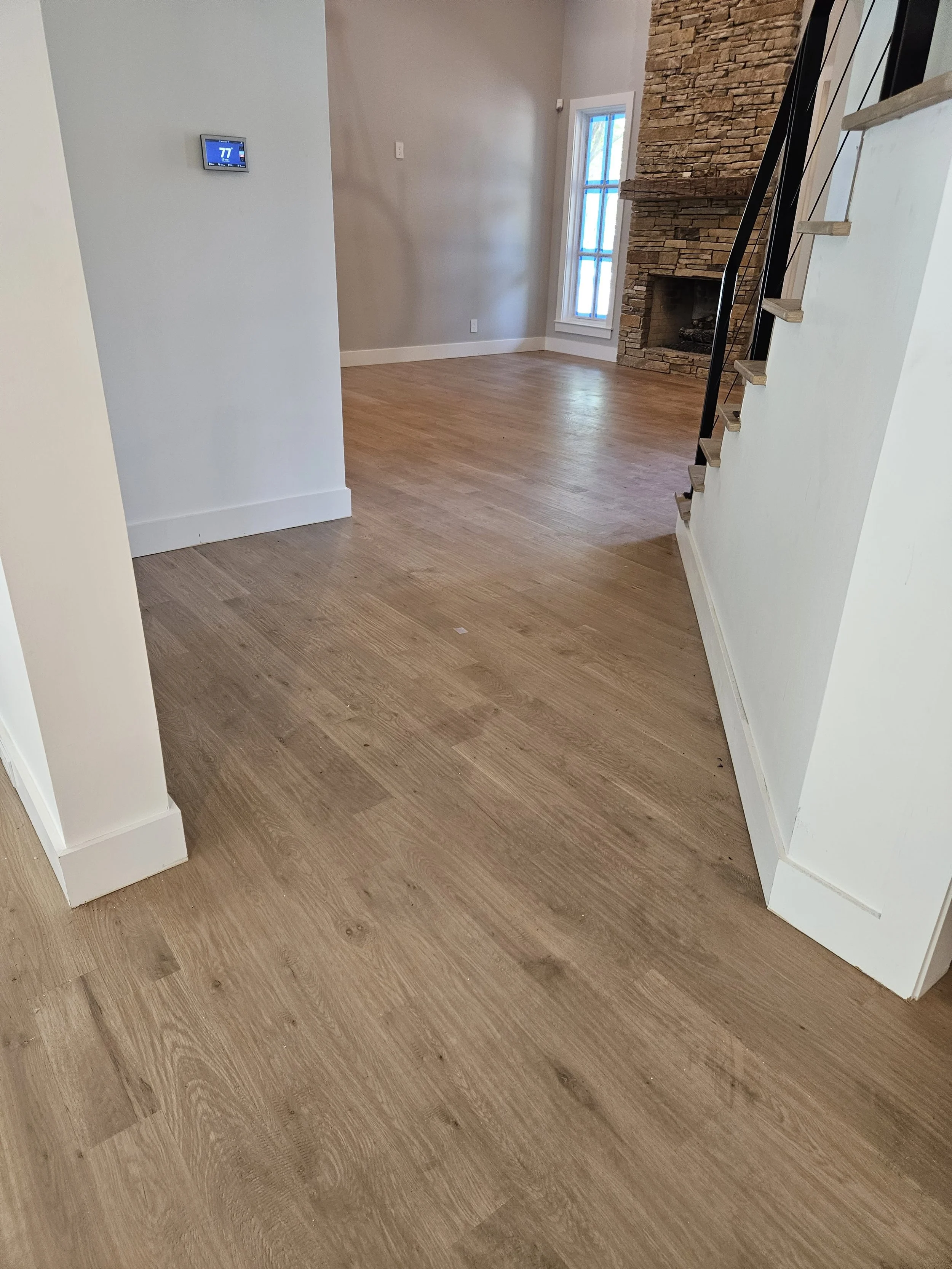Empty living room with hardwood floors, a white wall with a thermostat, a stone fireplace, a large window with blue grid pattern, and a staircase with wooden treads and black railing.