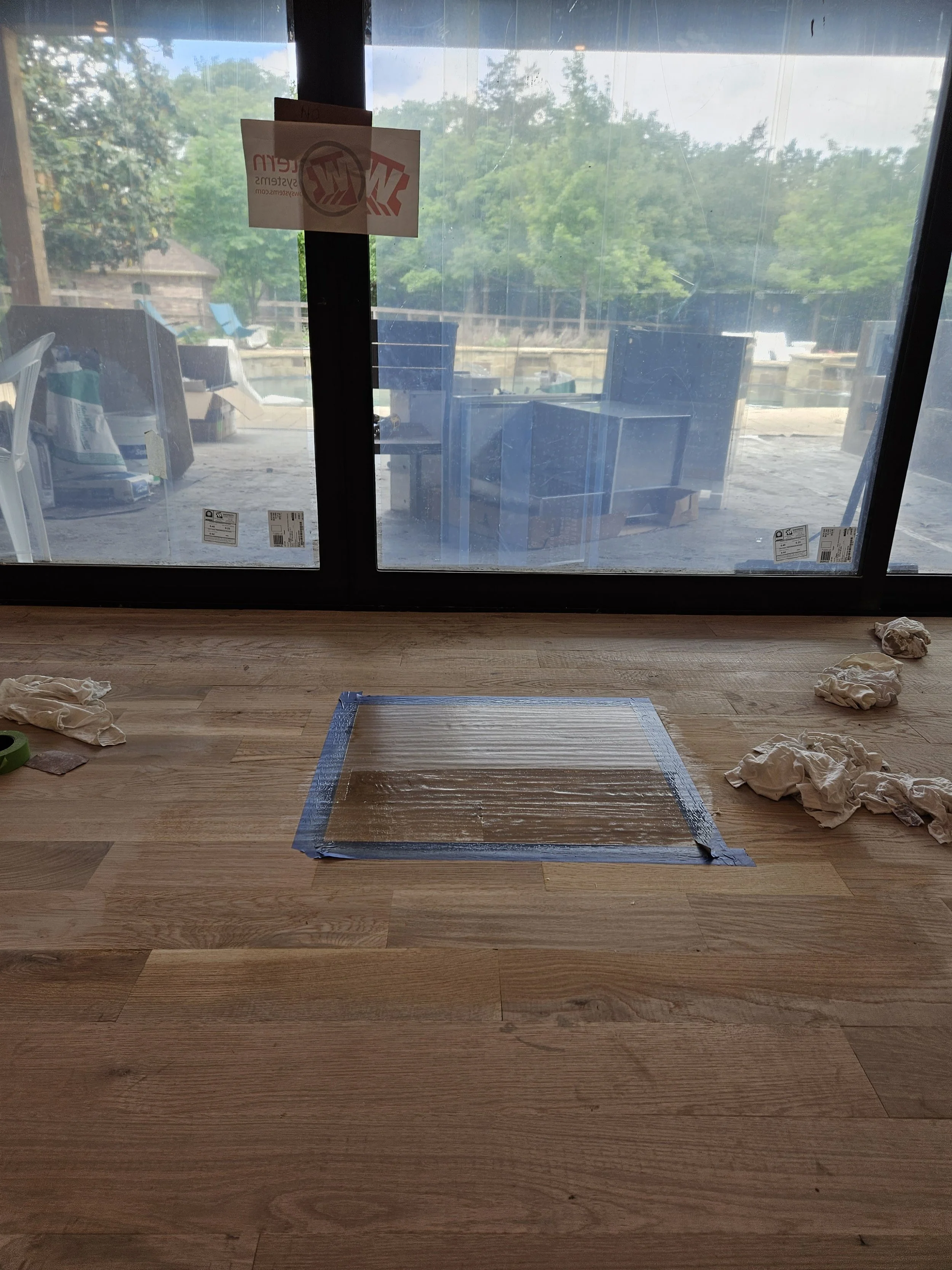 A wooden floor with a taped hole for a vent or air vent, several crumpled white cloths or rags scattered around, and a large glass door looking outside with outdoor furniture and trees visible.